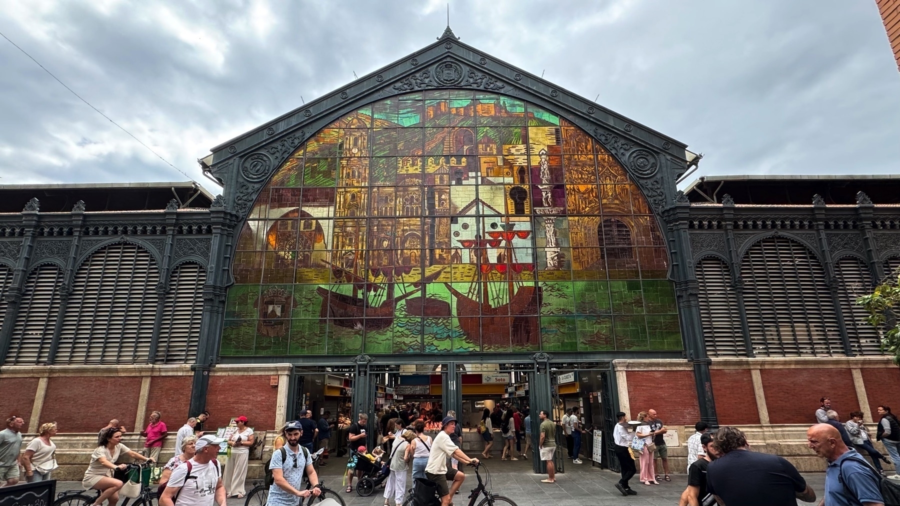 Front façade of the Atarazanas Market featuring a large, colorful stained-glass window depicting historical scenes, including ships and classical buildings. The window is set within a cast-iron frame of the market’s industrial architecture. A lively crowd of people walks, cycles, and shops in the foreground, adding energy and movement to the scene.