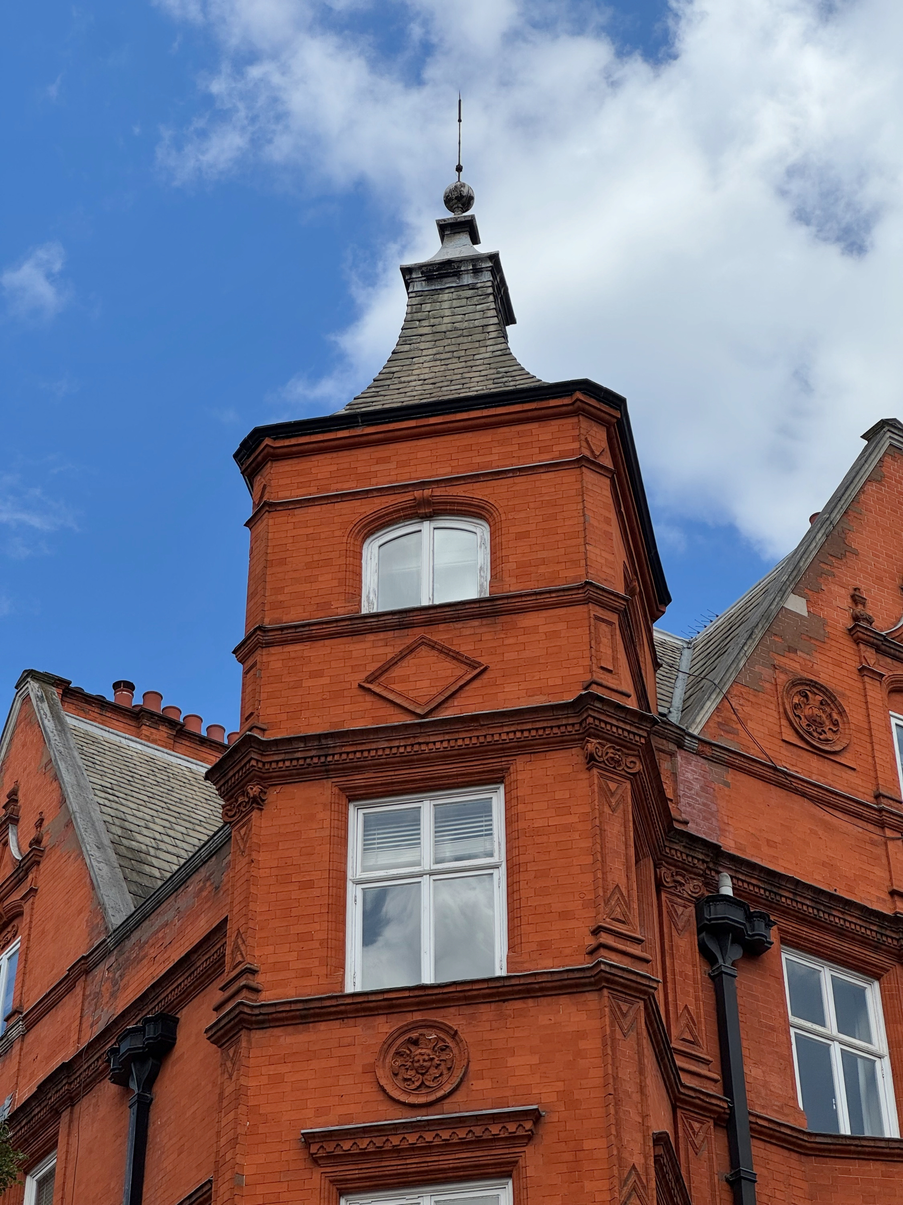 Ornate red-brick Victorian building with a corner tower, white sash windows, decorative stonework, and a steep slate roof topped by a finial against a bright blue sky with scattered clouds.