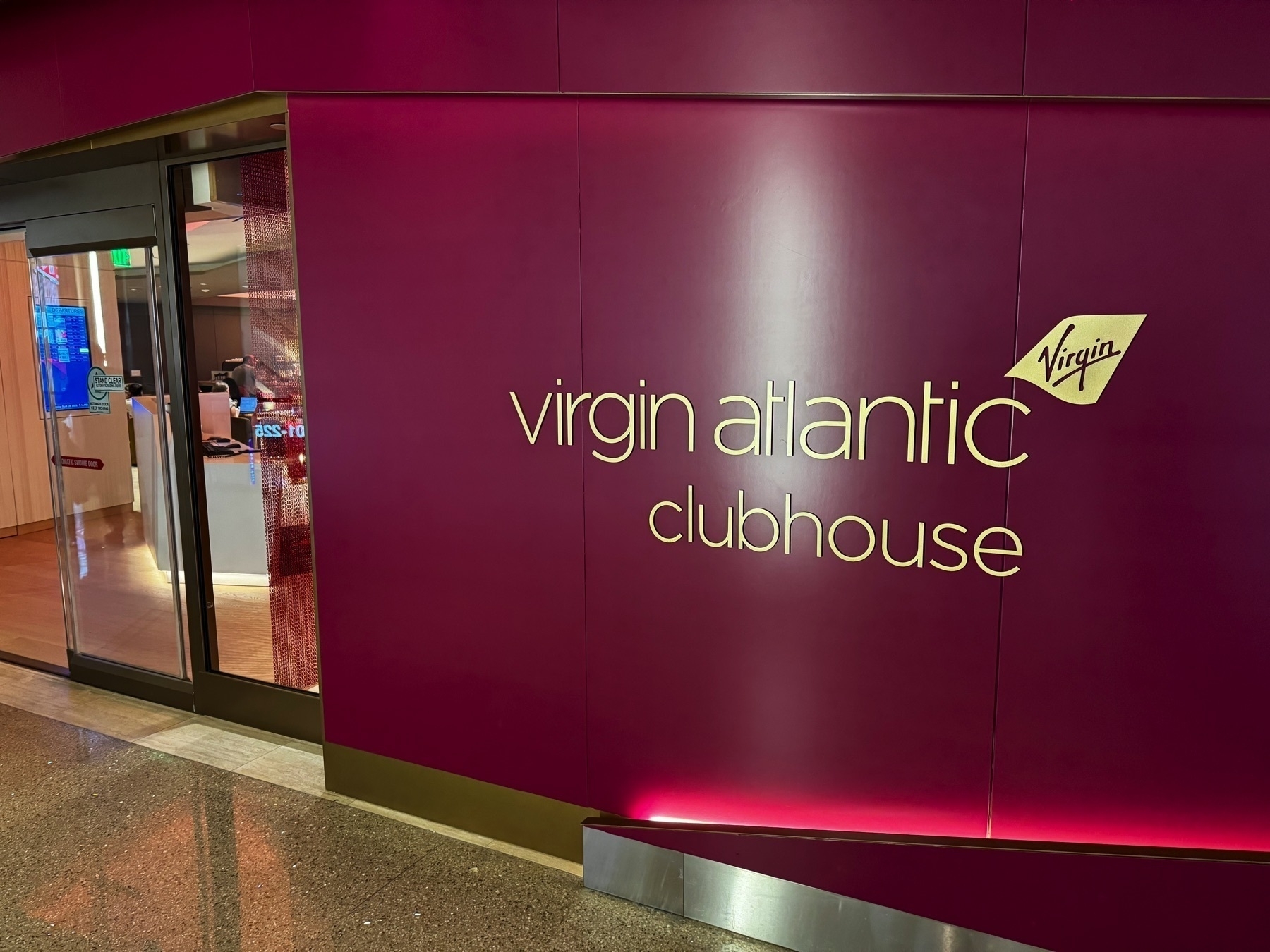 Entrance to the Virgin Atlantic Clubhouse at LAX, featuring a deep magenta wall with gold lettering and logo, and a glimpse of the sleek interior through the open glass door.