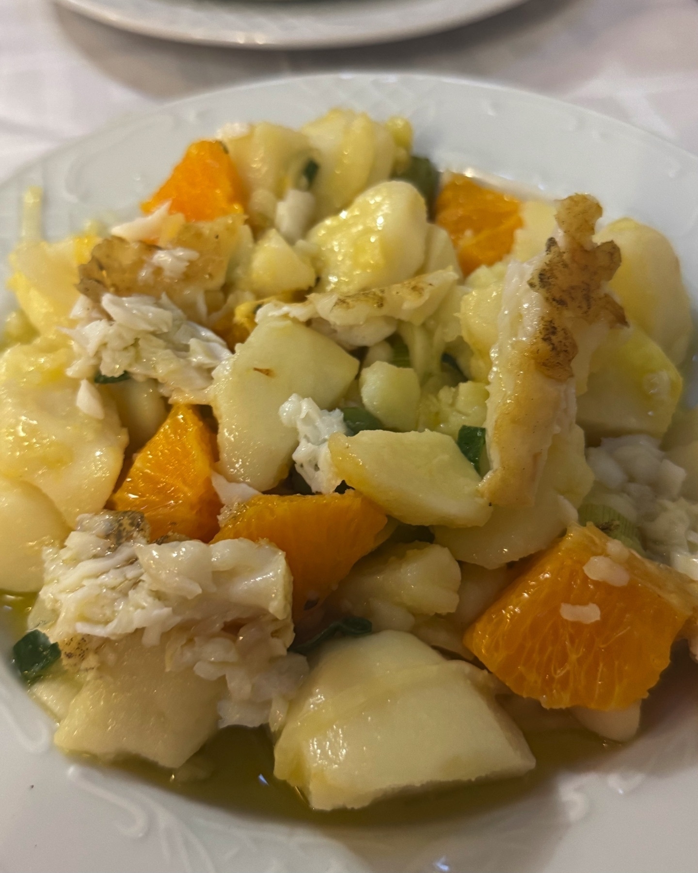A bright and refreshing cold salad composed of flaky salt cod, boiled potato chunks, and juicy orange wedges. Everything is lightly dressed with olive oil and garnished with sliced green onions, offering a mix of creamy, citrusy, and savory elements on a white plate.