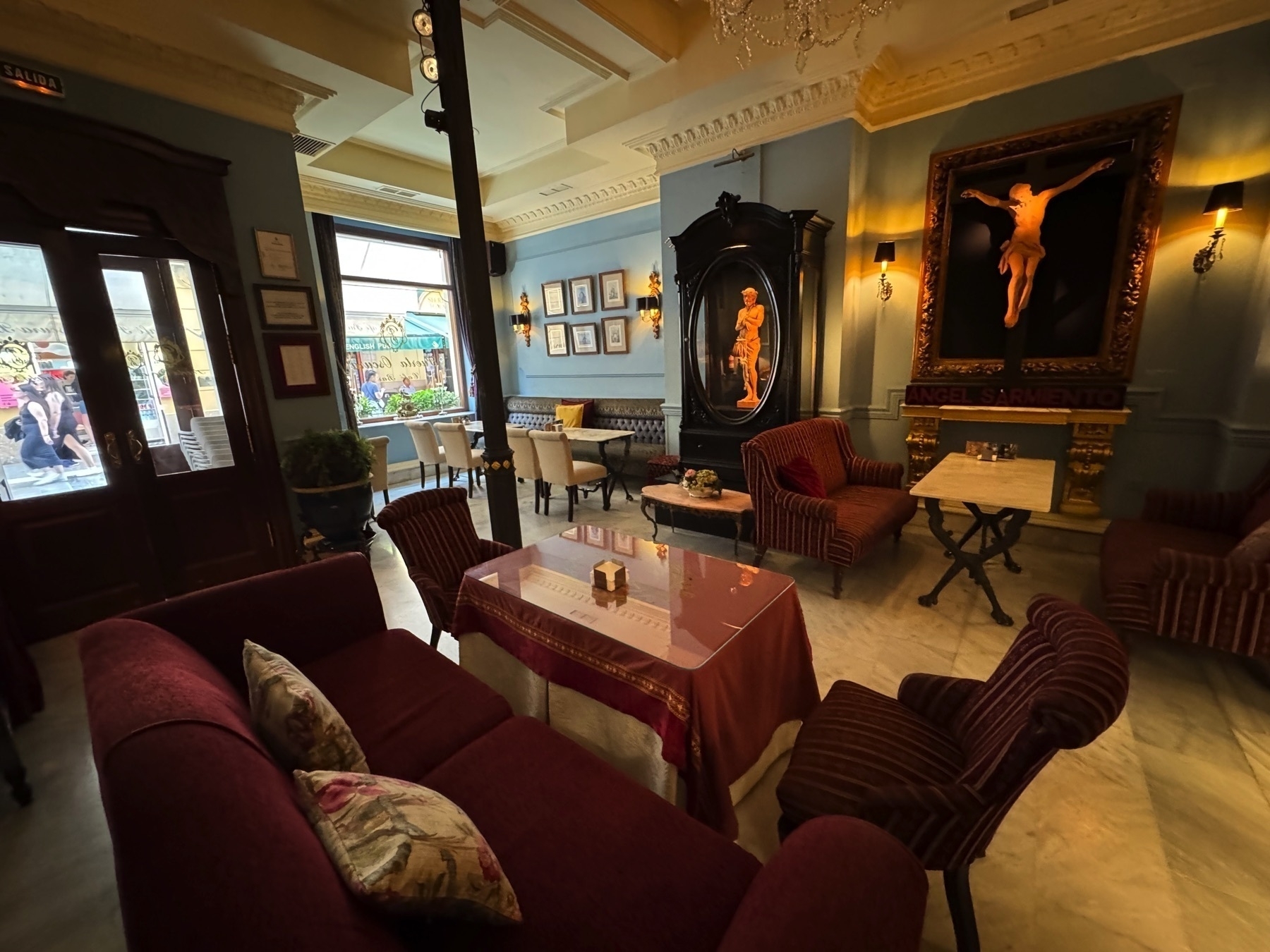 An elegant bar interior with plush velvet chairs, chandeliers, ornate frames, and religious art. The space combines vintage charm with a moody, intimate atmosphere perfect for a relaxed evening drink.