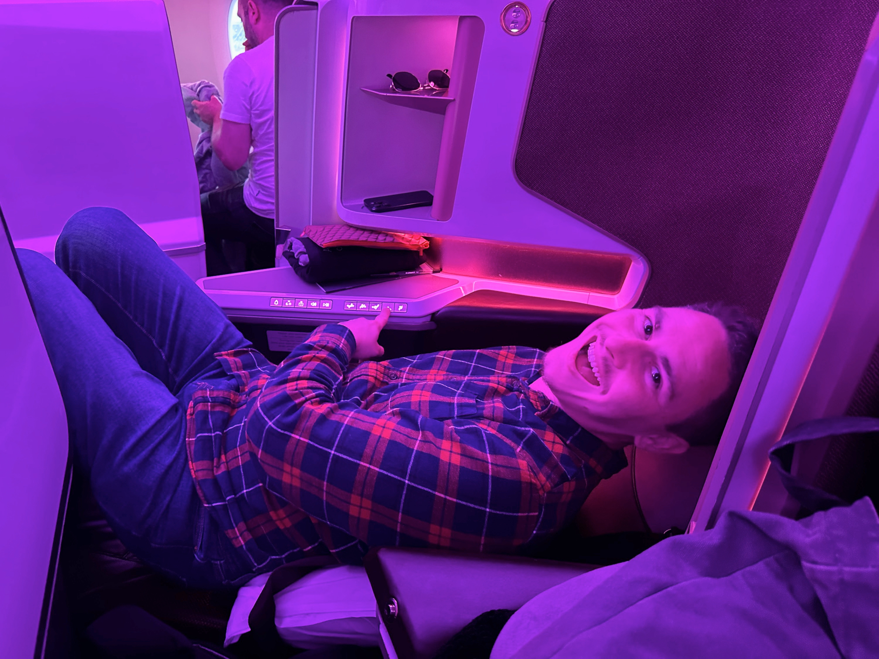Me reclining and grinning in a spacious business class airplane seat under purple cabin lighting, pointing at the seat controls with personal storage and another passenger visible nearby.