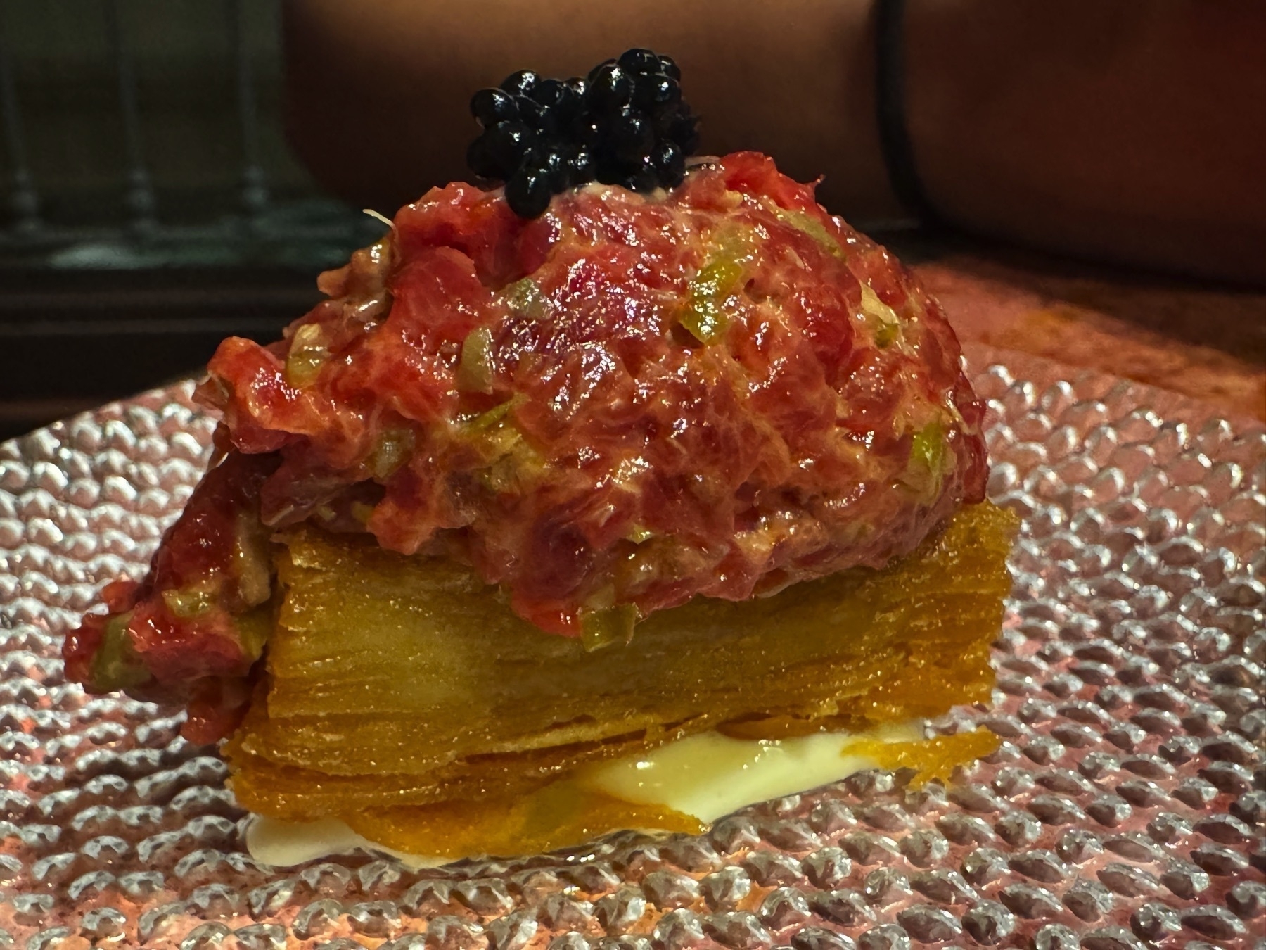 A composed dish featuring a golden potato mille-feuille topped with finely chopped beef tartare, mixed with pickles or green herbs, and finished with a dollop of glossy black caviar. The dish sits on a glass plate, with a layer of creamy sauce beneath the potato adding contrast.