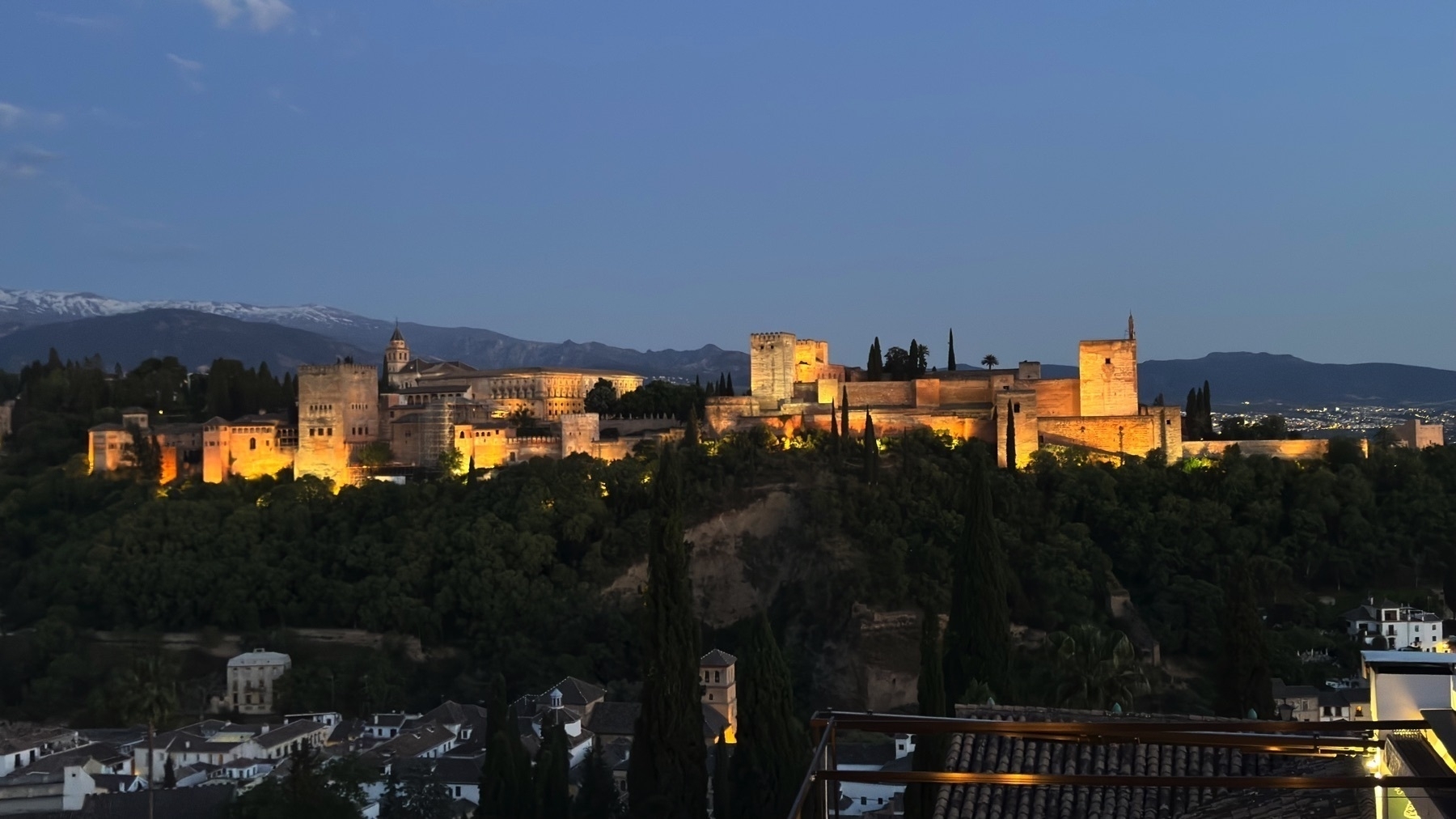 Panoramic twilight view of the Alhambra glowing under floodlights, with towers and walls sharply defined against a darkening sky. The rooftops of the Albaicín and several terrace structures are visible in the foreground, adding a sense of depth and scale.