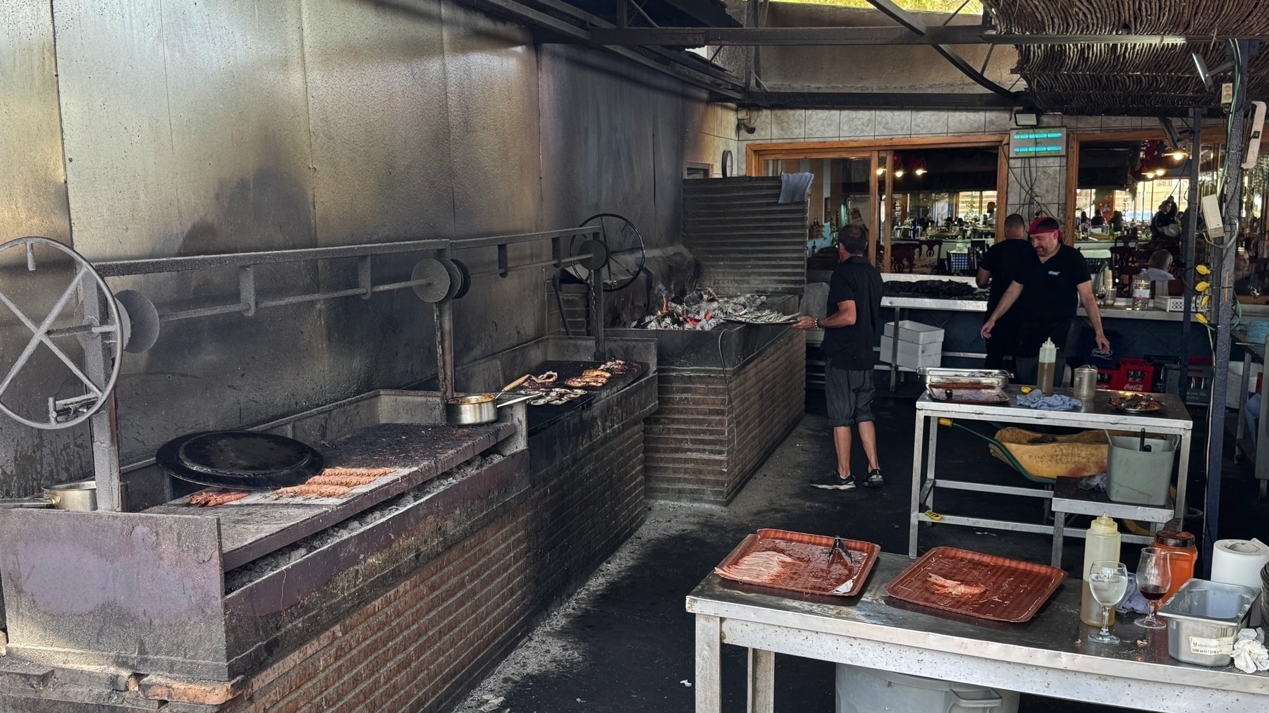 Industrial-style open kitchen with large charcoal grills and trays of food in preparation. Cooks in black uniforms work near the smoky grills, with skewers and meats visibly charring over hot coals.