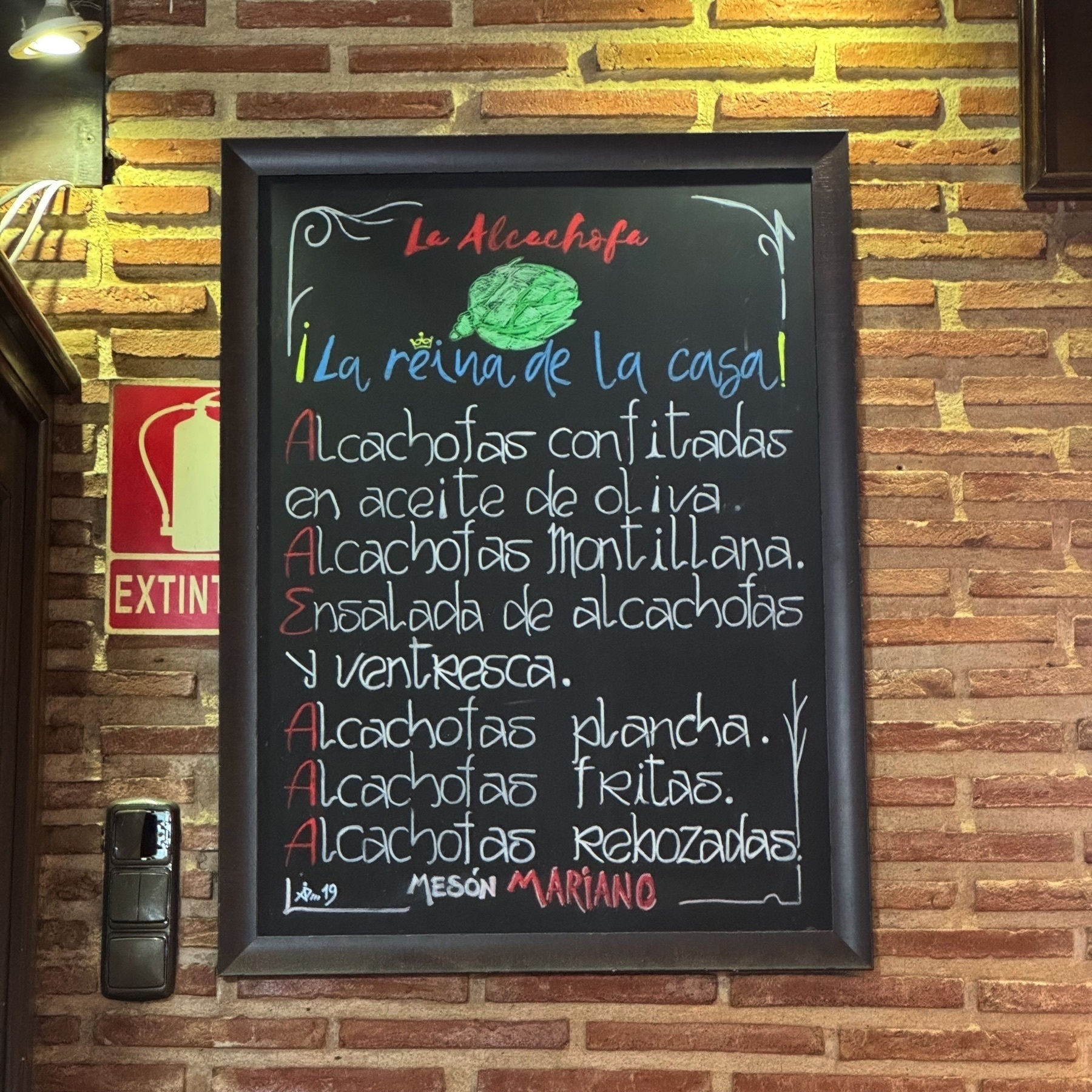 A chalkboard menu on a brick wall titled “La Alcachofa” (The Artichoke), featuring a list of house specialties centered around artichokes—confited, Montillana-style, salad with tuna belly, grilled, fried, and battered. The vibrant chalk colors and hand-drawn artichoke illustration give a cozy, welcoming feel.