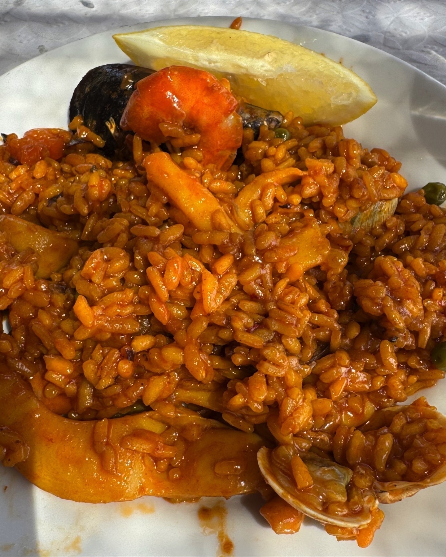 Hearty serving of seafood paella featuring saffron-tinted rice mixed with mussels, shrimp, squid, and vegetables. A lemon wedge sits on the rim of the plate.