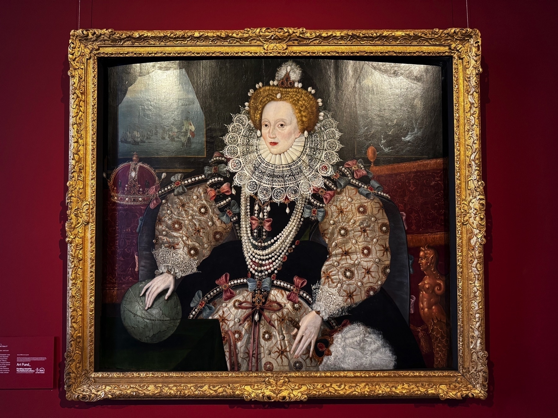 The iconic Armada Portrait of Queen Elizabeth I, displayed against a rich red wall and surrounded by a gilded frame. The queen wears an elaborate gown adorned with pearls and embroidery, one hand resting on a globe and the other near a chair. Her attire and posture emphasize power and wealth. Behind her, the backdrop shows scenes of the Spanish Armada’s defeat, underscoring the historic moment.