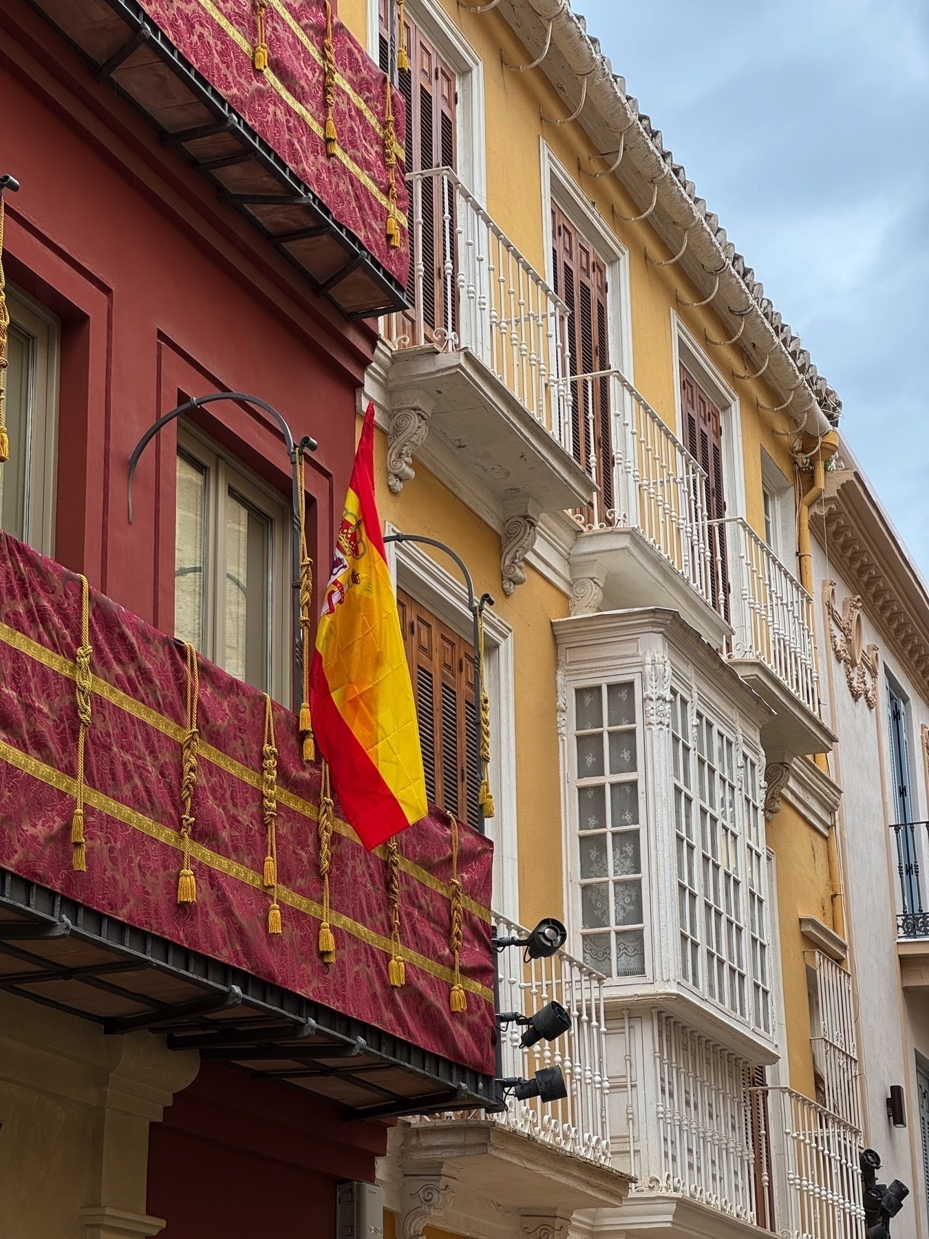 Decorated balconies in Málaga’s city center, featuring maroon velvet fabric with gold tassels and a Spanish flag with a detailed coat of arms hanging from a wrought-iron bar.