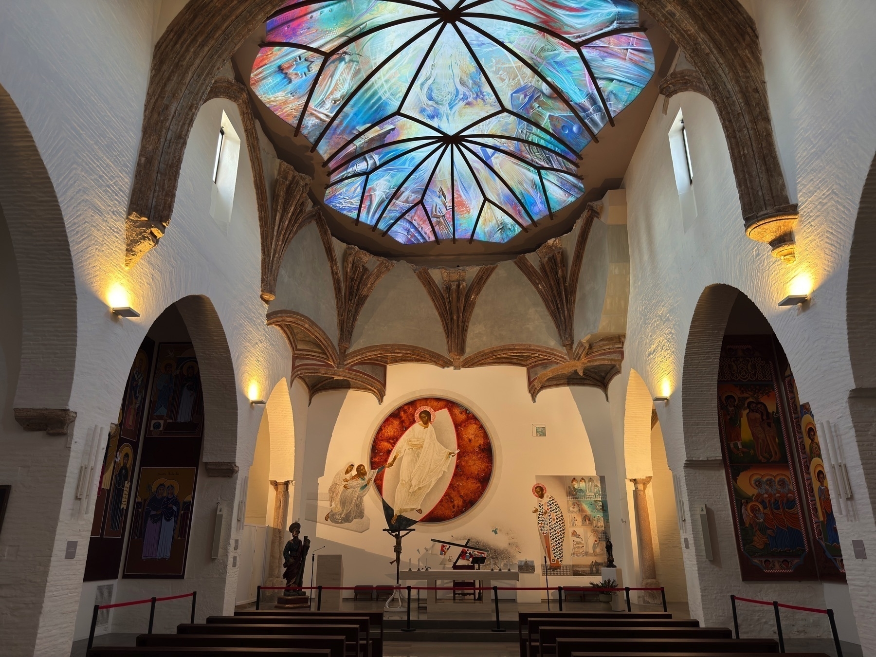 Interior of a chapel with a vivid, modern stained-glass ceiling in swirling colors, set above ribbed Gothic-style arches. Below, religious artwork and sculptures are displayed near an altar, blending traditional and contemporary elements.