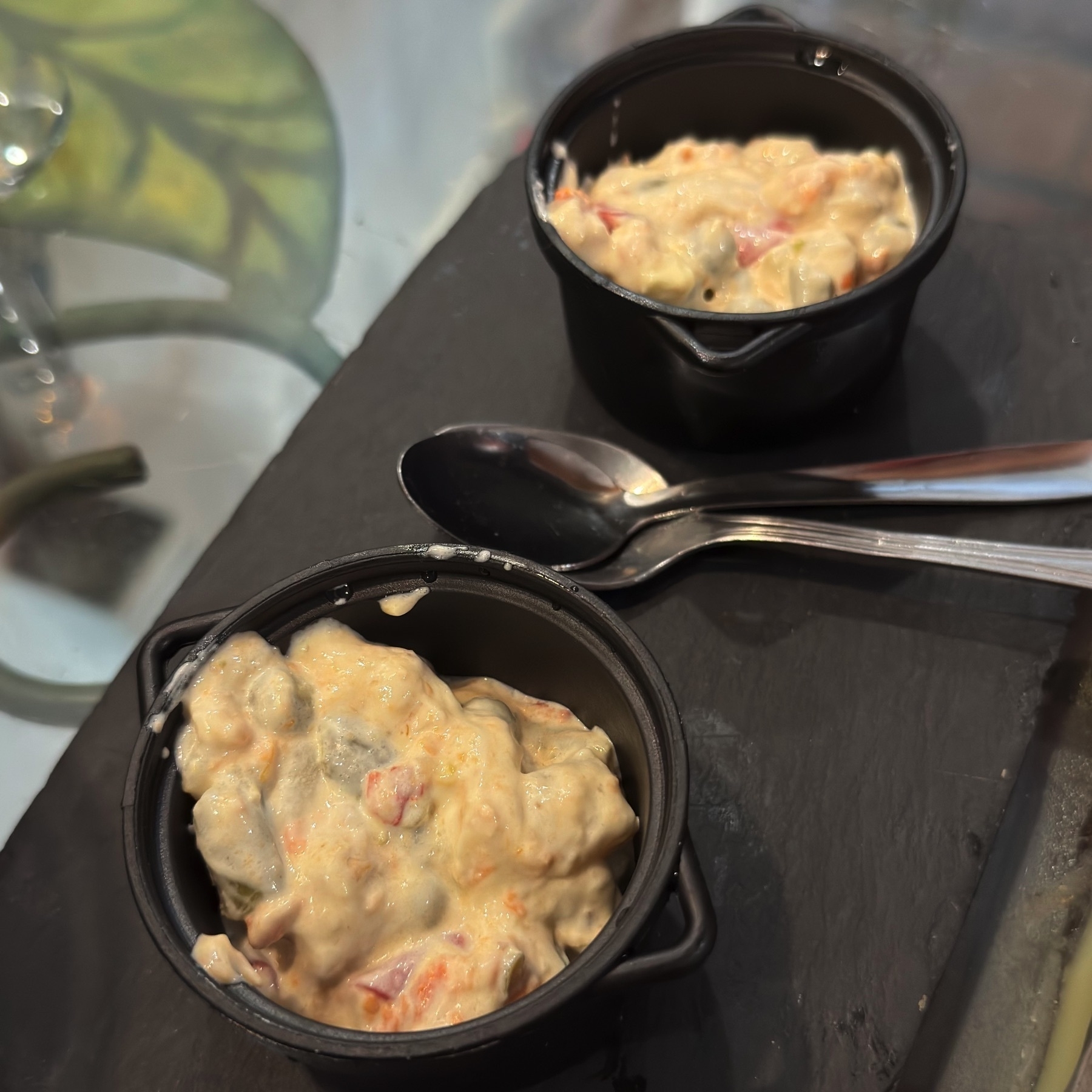 Small cast-iron pots filled with a creamy potato and tuna salad, speckled with diced vegetables such as red pepper and pickles. Served on a slate board with metal spoons, the rustic presentation highlights a traditional tapa with comforting, chilled textures.