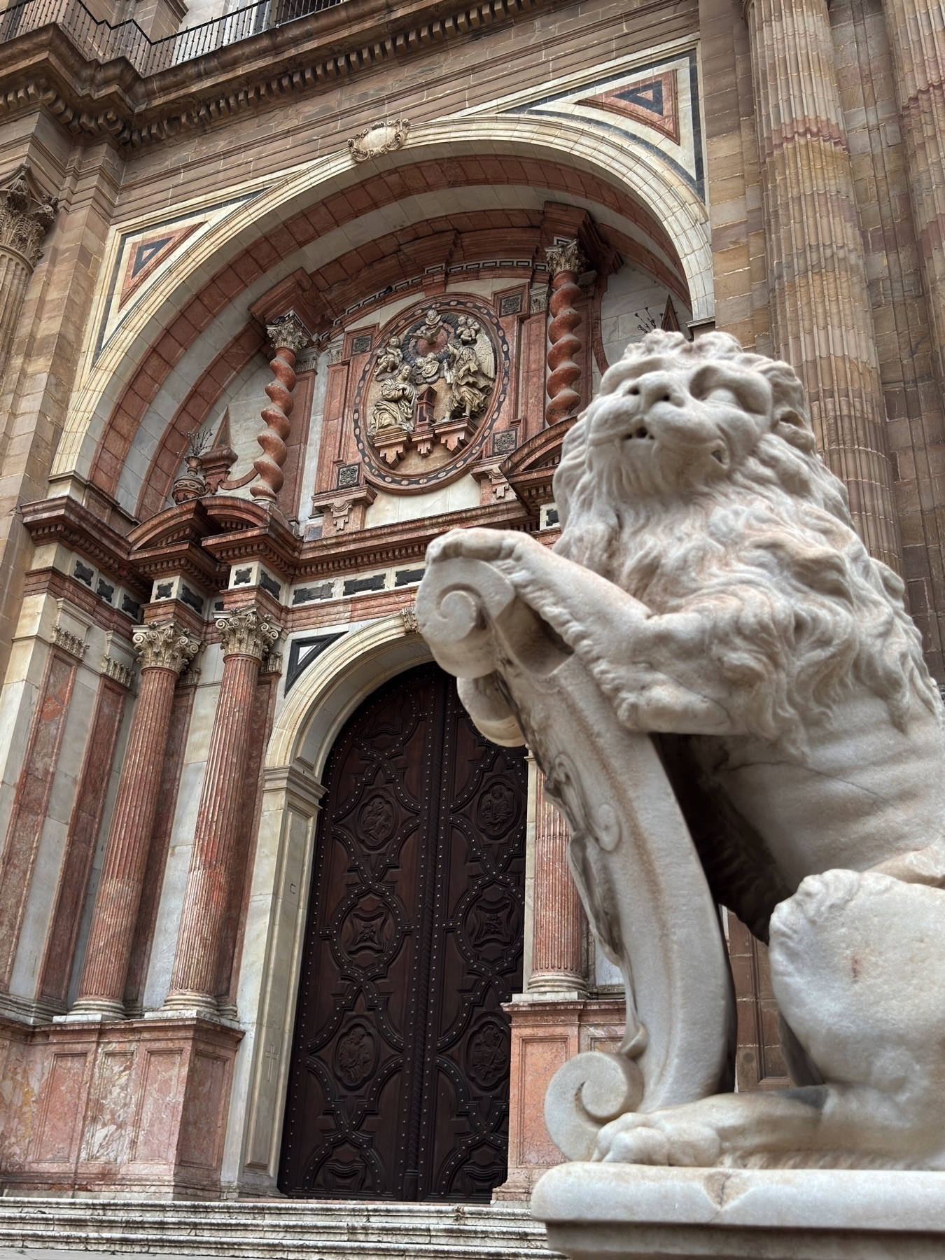 Close-up of a richly decorated entrance to the Málaga Cathedral, featuring a marble lion statue in the foreground holding a shield. Behind it, the grand wooden doors are framed by intricate stonework, twisted columns, and a colorful coat of arms with religious figures carved in relief. The composition emphasizes the detail and craftsmanship of the Baroque façade.