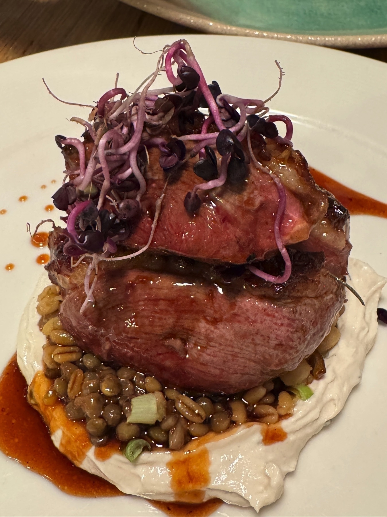Seared duck medallion stacked over a bed of grains and creamy purée, topped with purple microgreens and finished with a glossy reduction, blending rustic and refined elements.