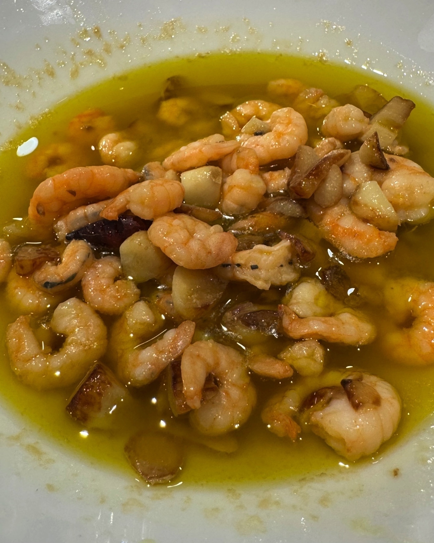 A bowl of sizzling gambas al ajillo—plump shrimp and diced garlic swimming in hot olive oil. The prawns are pink and slightly curled, with golden garlic bits adding texture and a savory aroma. The oil’s sheen reflects the warmth and flavor of the dish.