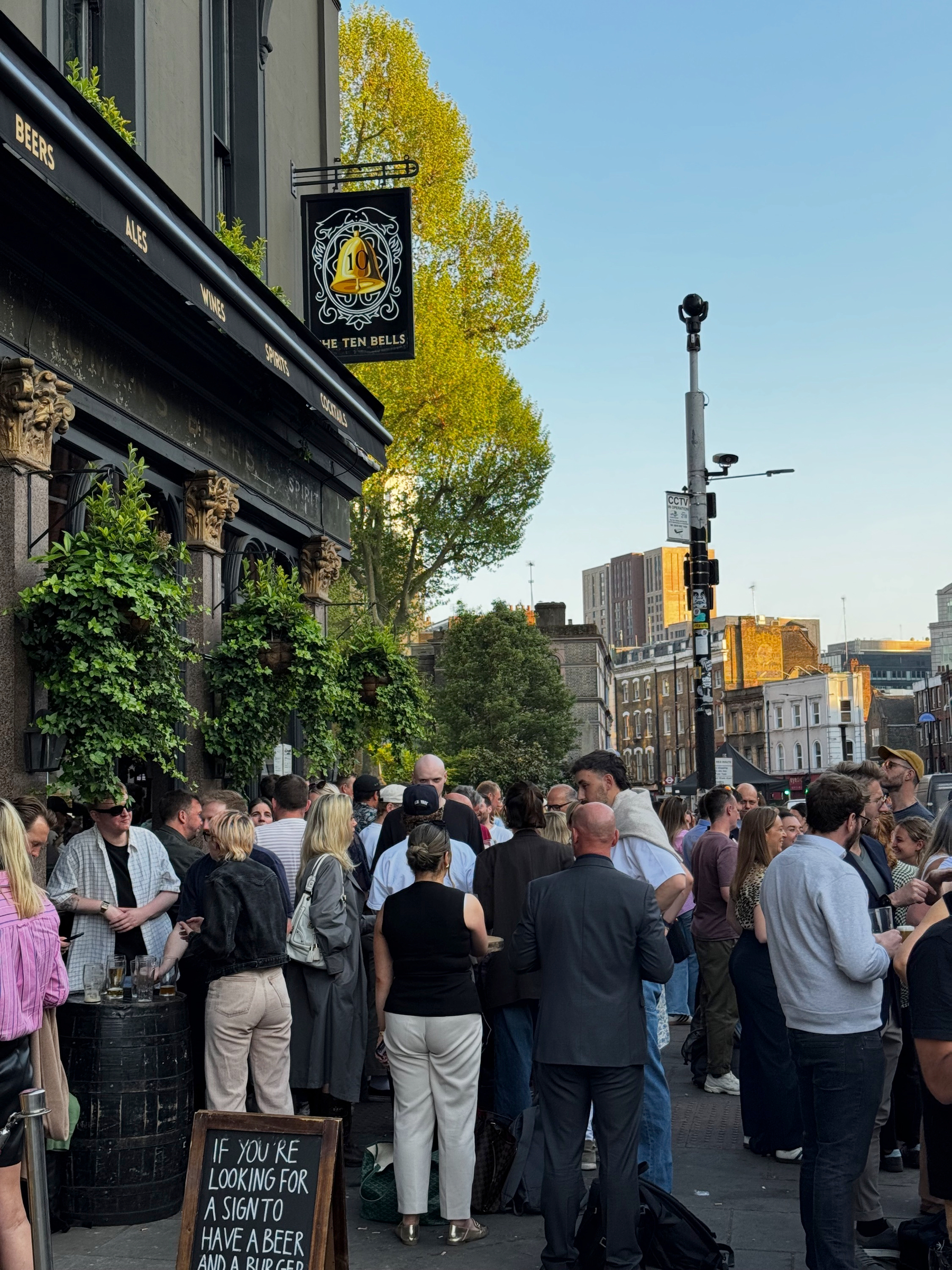 Crowd gathered outside The Ten Bells pub in Spitalfields, with people holding drinks beneath the hanging pub sign, leafy greenery on the facade, and a busy street atmosphere.