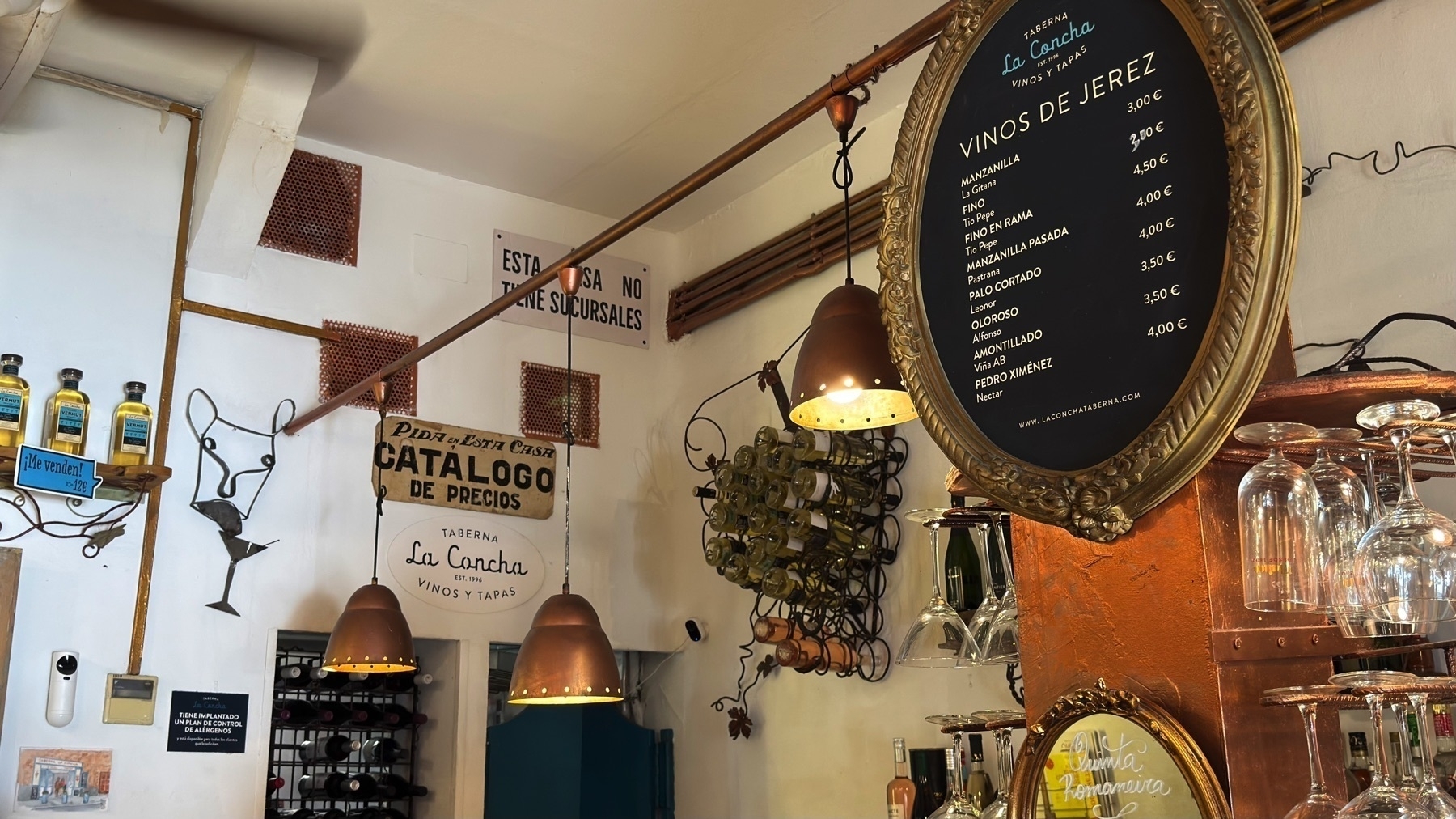 Interior shot of the wine and bar area at Taberna La Concha, featuring a gold-framed chalkboard listing Jerez wines, a wall-mounted rack of white wine bottles, and rows of glassware hanging from copper shelving. Warm, vintage-style pendant lights and layered signage contribute to a cozy and curated atmosphere.