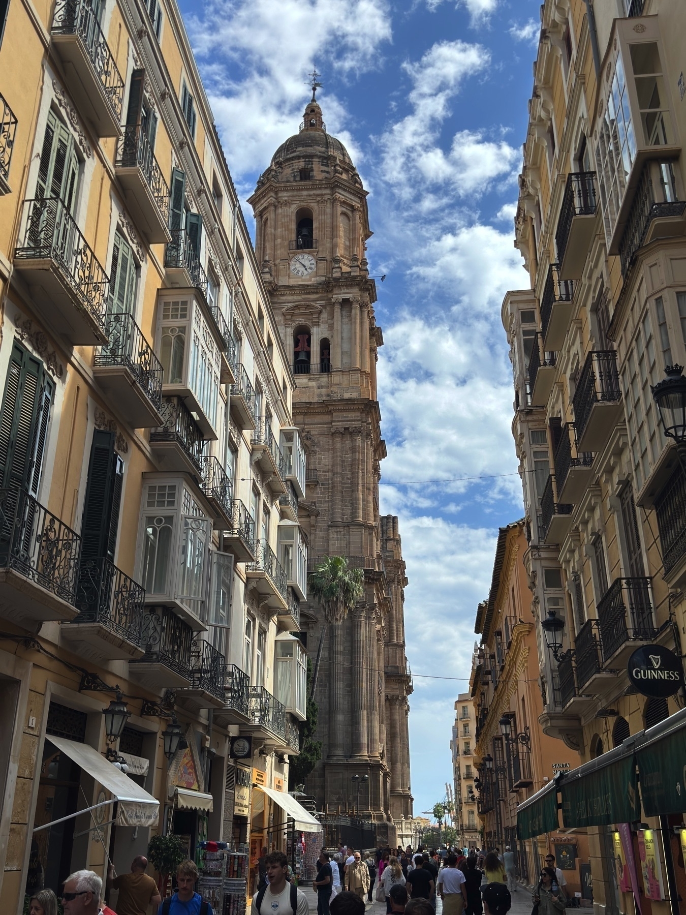 Crowded street scene with the cathedral towering between rows of historic apartment buildings. Open balconies, shop signs, and a partly cloudy sky frame the Gothic-Renaissance cathedral.