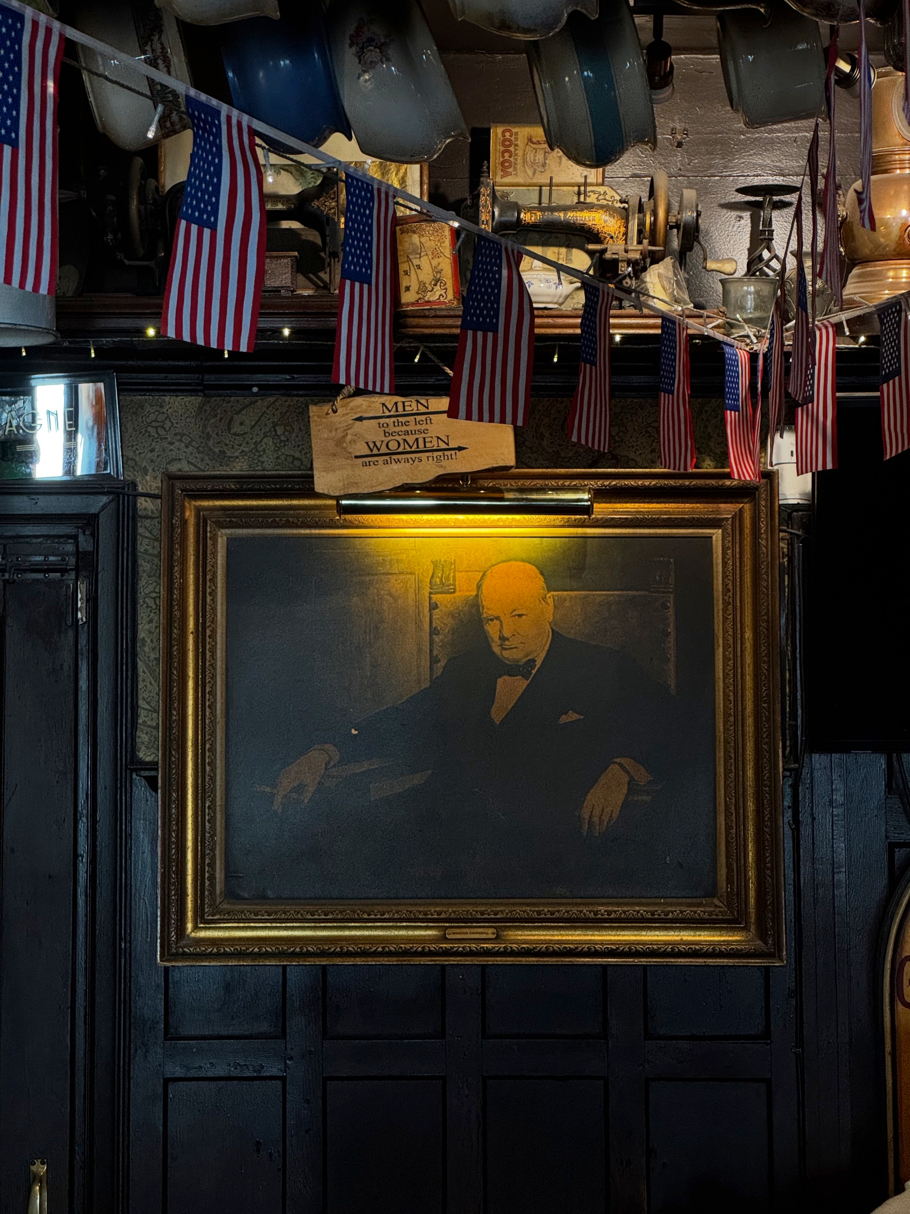 Framed portrait of Winston Churchill hanging on dark wood paneling beneath small American flags and a humorous sign for the restroom: “Men to the left because women are always right!”