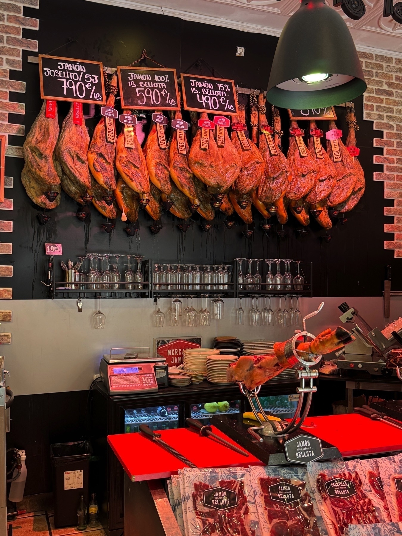 An interior view of a jamón shop with multiple legs of cured ham hanging on black hooks behind the counter, each tagged with red and black labels reading “Bellota.” Chalkboard signs display different types and prices of jamón per kilo, including 100% Ibérico and 75% Ibérico. A carving station in the foreground features a mounted ham leg and slicing knives, with vacuum-sealed packs of jamón and glassware neatly arranged below.