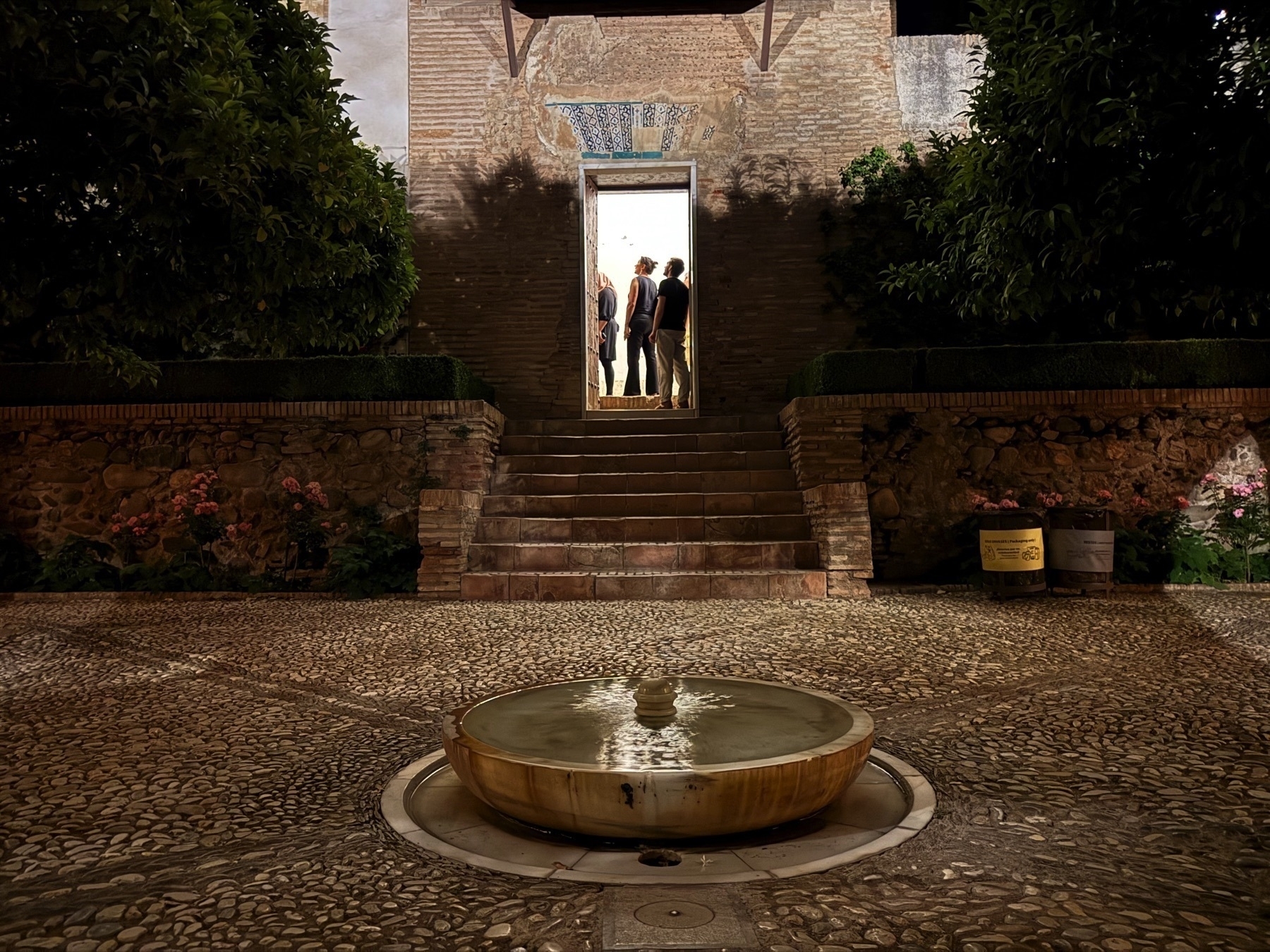 A quiet courtyard near the Generalife, with a small circular stone fountain in the foreground and a softly lit staircase leading to a doorway where a group of visitors stands under a warm light.