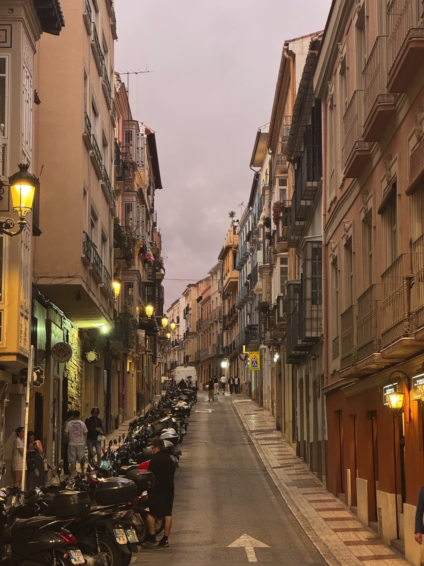 Twilight scene on Calle Carretería, a narrow uphill street lined with scooters, pedestrians, and traditional buildings with balconies and lit lanterns, capturing the peaceful evening atmosphere in the historic center.