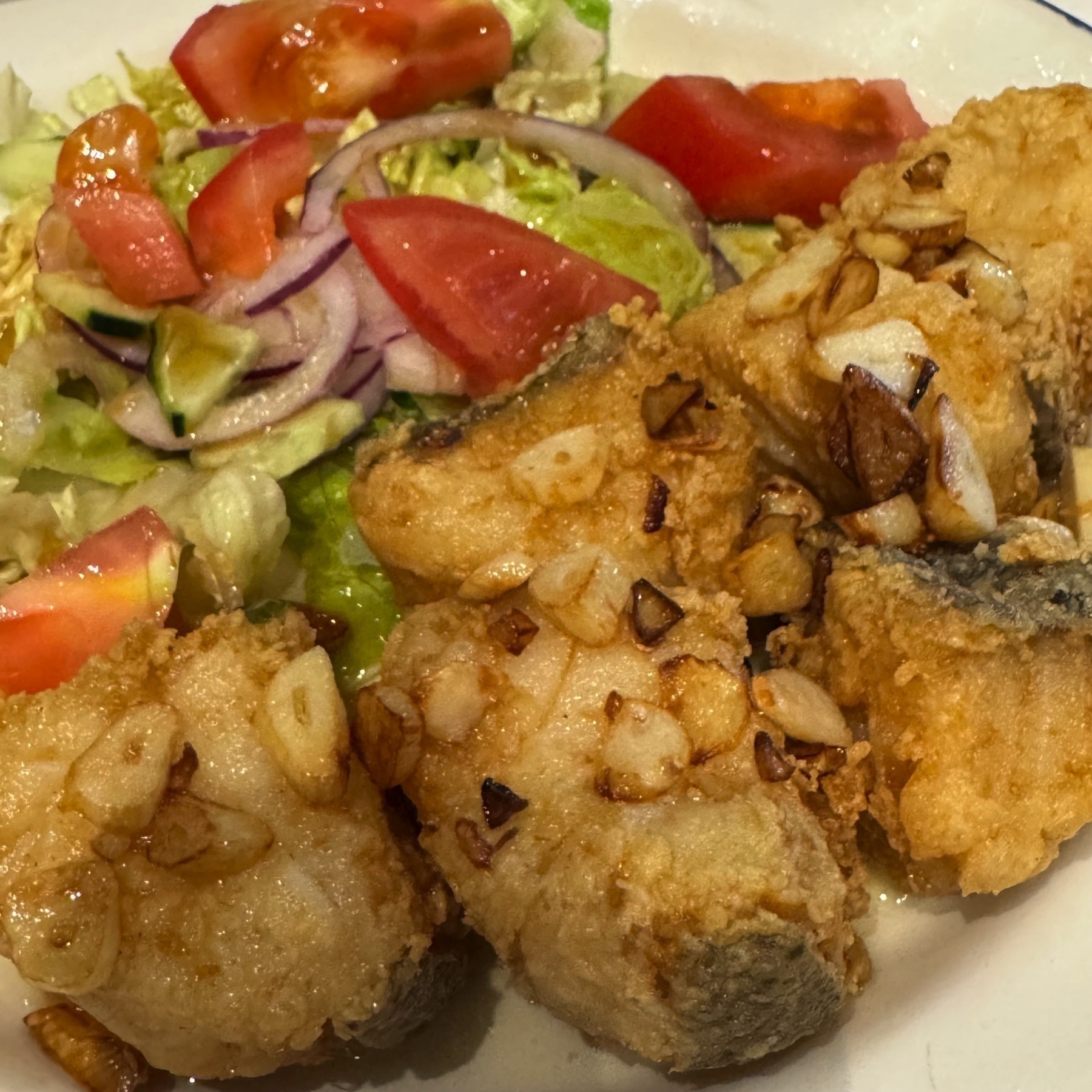 Golden, lightly battered cod pieces topped with crispy slivers of garlic, served alongside a fresh tomato and lettuce salad with red onion. The contrast between the crunchy garlic, juicy vegetables, and flaky fish makes the dish look satisfying and well-balanced.