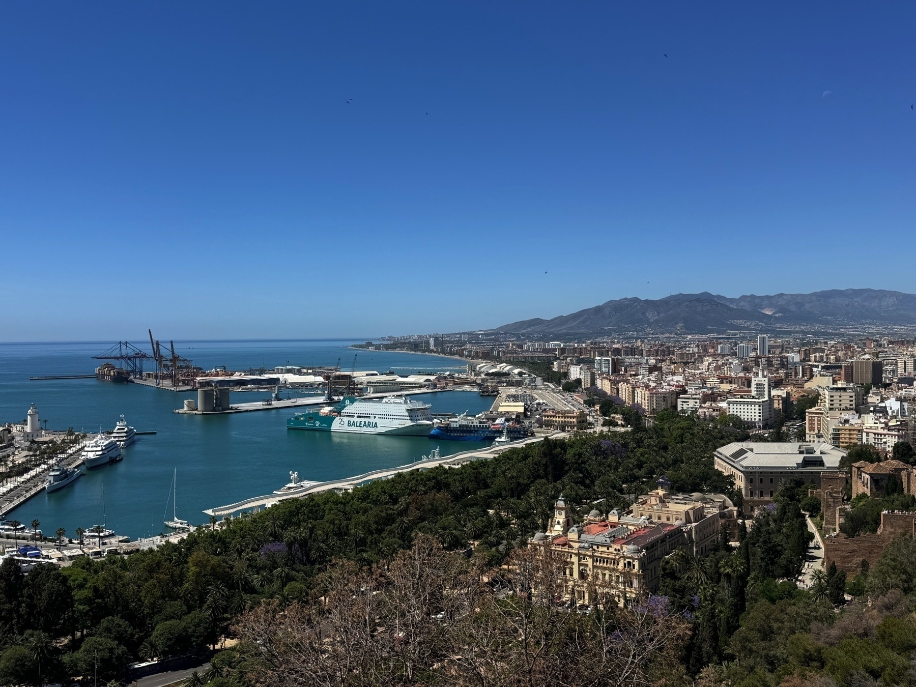 Elevated view of Málaga’s port area with deep turquoise waters, yachts, ferries, and shipping cranes. The city’s architecture extends inland toward mountains, with the lush greenery of parkland in the foreground.