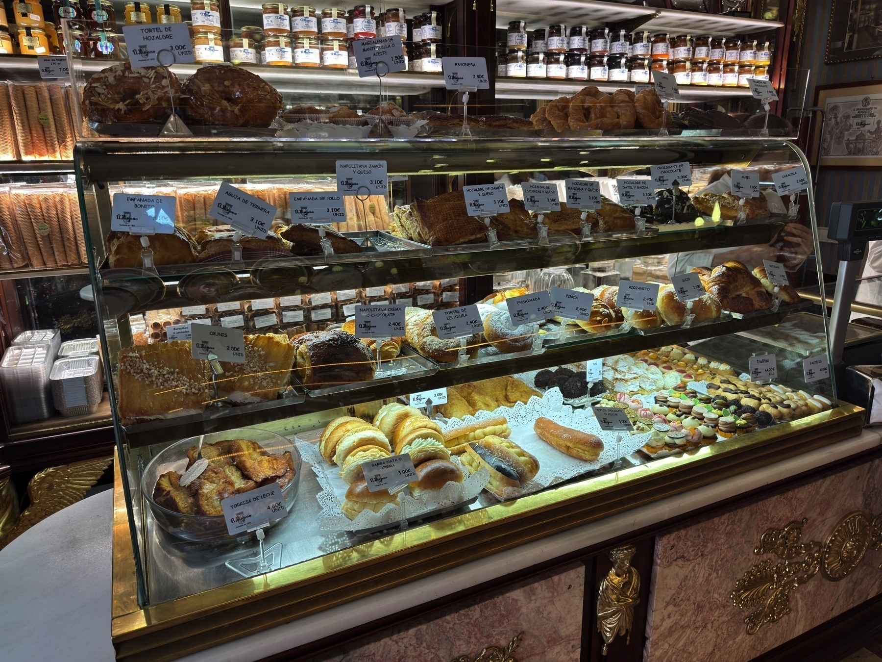 A well-lit glass pastry display brims with traditional Spanish baked goods arranged across three shelves. Each level features neatly labeled treats such as empanadas, croissants, napolitanas, puff pastries, and cookies, framed by a backdrop of decorative jars and an opulent wooden and brass case. The abundance and variety—ranging from flaky to golden to sugar-dusted—create a tempting and indulgent visual feast.