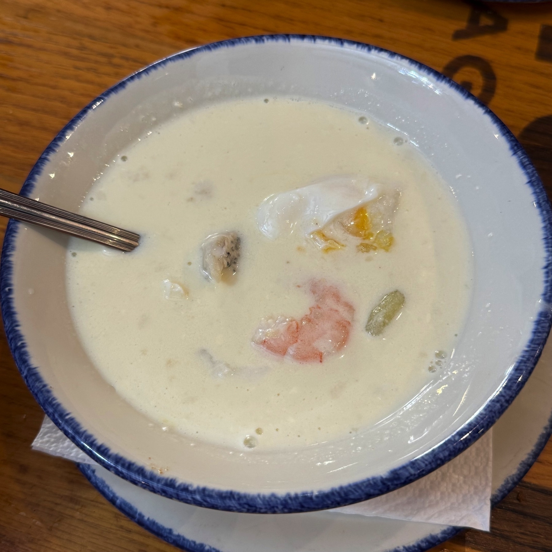 A bowl of thick, white soup with visible pieces of seafood—shrimp, white fish, and poached egg—floating in the rich, velvety base. The edges of the bowl are slightly rimmed with soup foam, indicating a warm, freshly served dish.