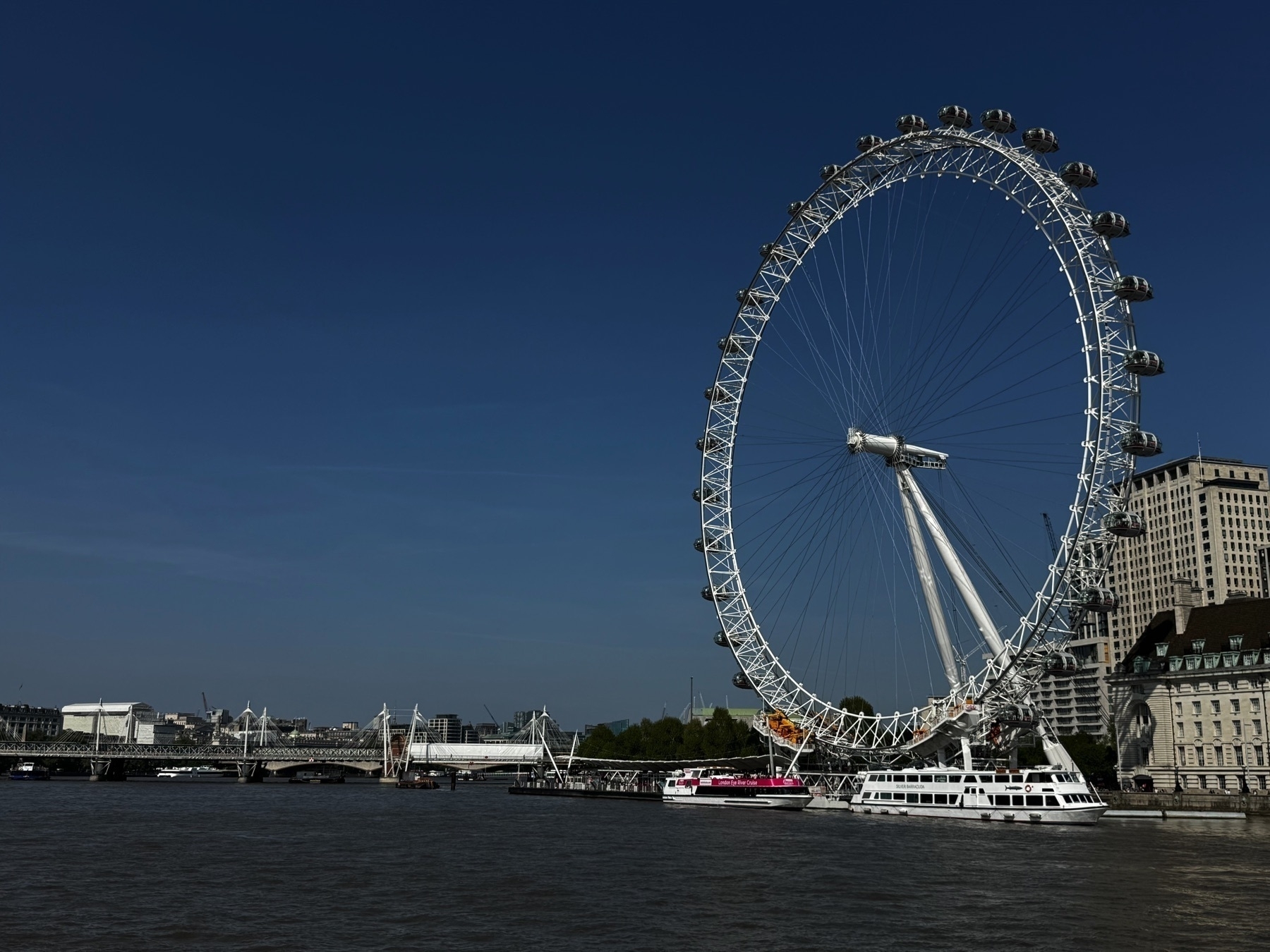 The London Eye seen from a distance across the River Thames, its full circular structure set against a deep blue sky. White boats are moored nearby, and a pedestrian bridge extends across the river to the left.