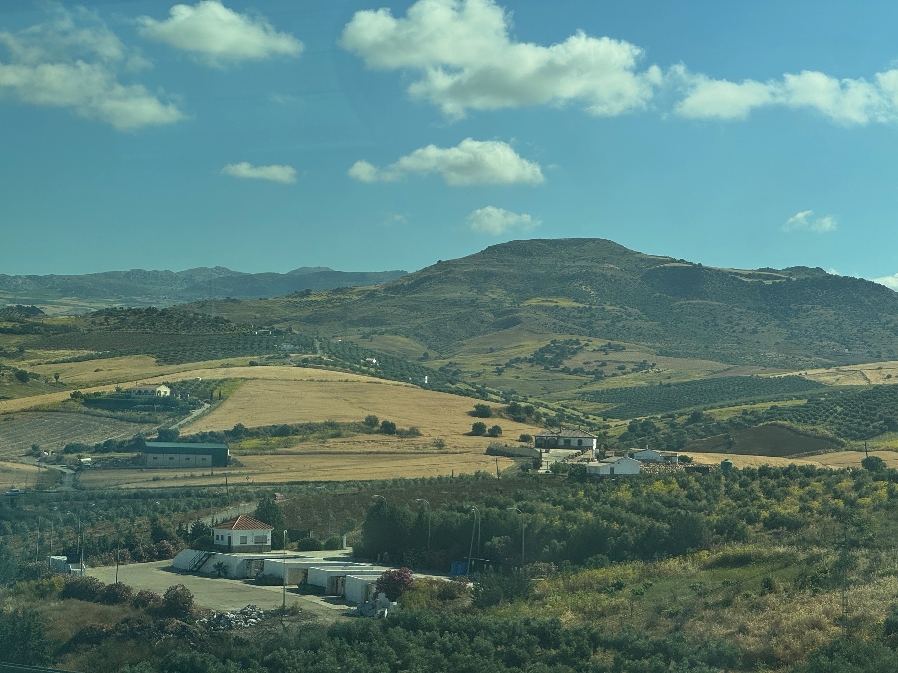 A scenic patchwork of cultivated fields and olive groves spreads across a valley surrounded by gently sloping mountains. Clusters of white buildings with terracotta roofs dot the landscape, with winding roads connecting them. The late afternoon sun casts warm light and long shadows, enhancing the contrast between the golden fields and green vegetation under a vibrant sky with scattered clouds.