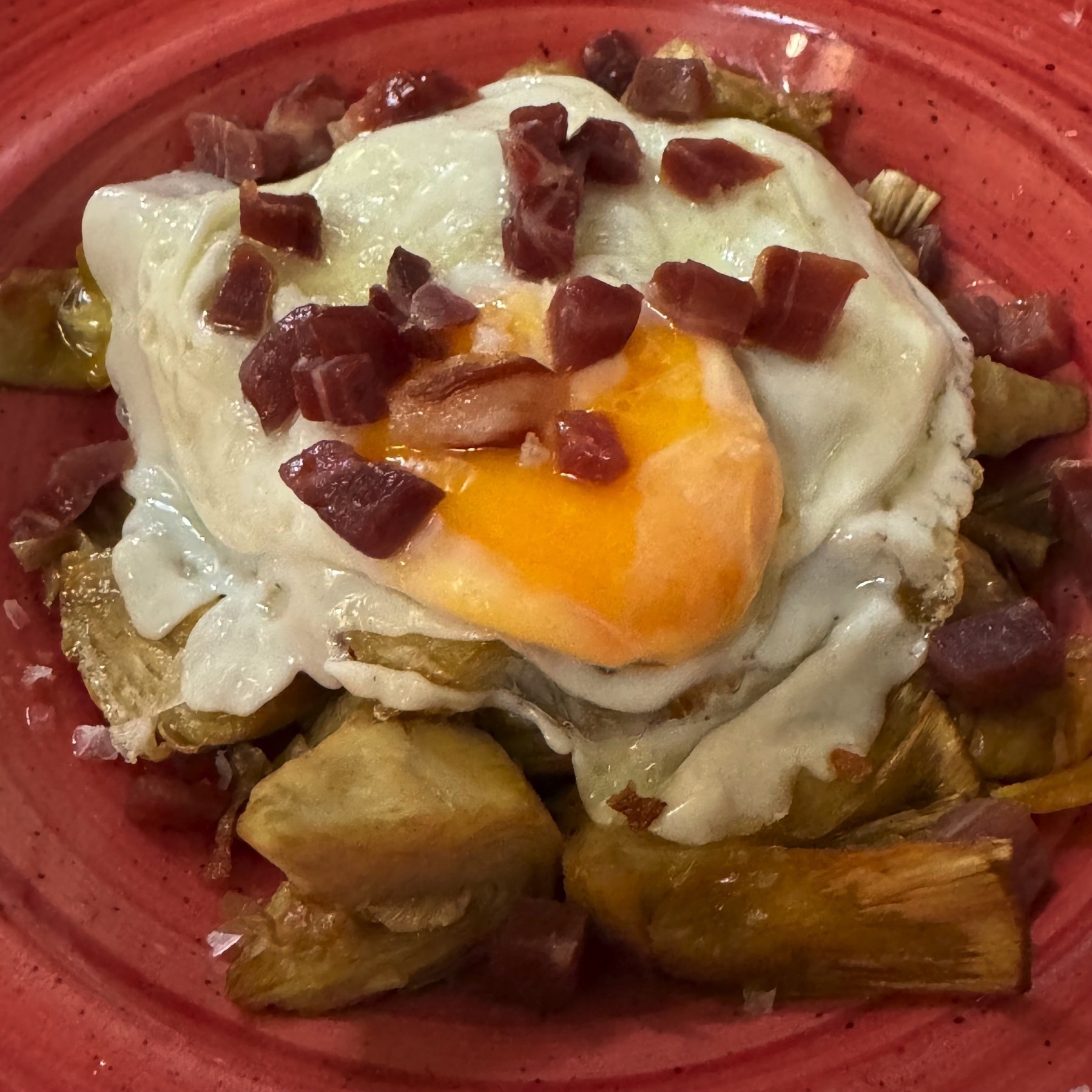 A hearty plate featuring a crispy-edged fried egg with a soft, runny yolk placed over a generous layer of golden-brown artichoke hearts. The dish is finished with a scattering of finely diced cured ham, adding rich color and texture. Presented on a rustic red ceramic plate that enhances its warmth and visual appeal.