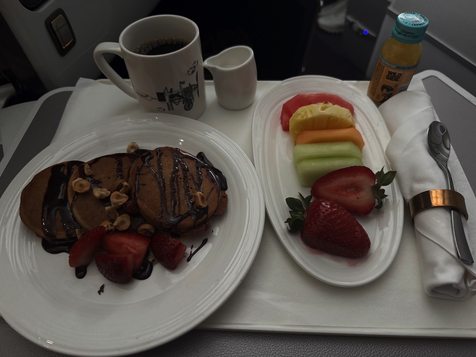 Breakfast tray with chocolate-drizzled pancakes topped with strawberries and hazelnuts on one plate, and a colorful fruit medley on another. Served with coffee, a small pitcher of cream, and a bottle of cold-pressed juice, all arranged neatly on a white tray.