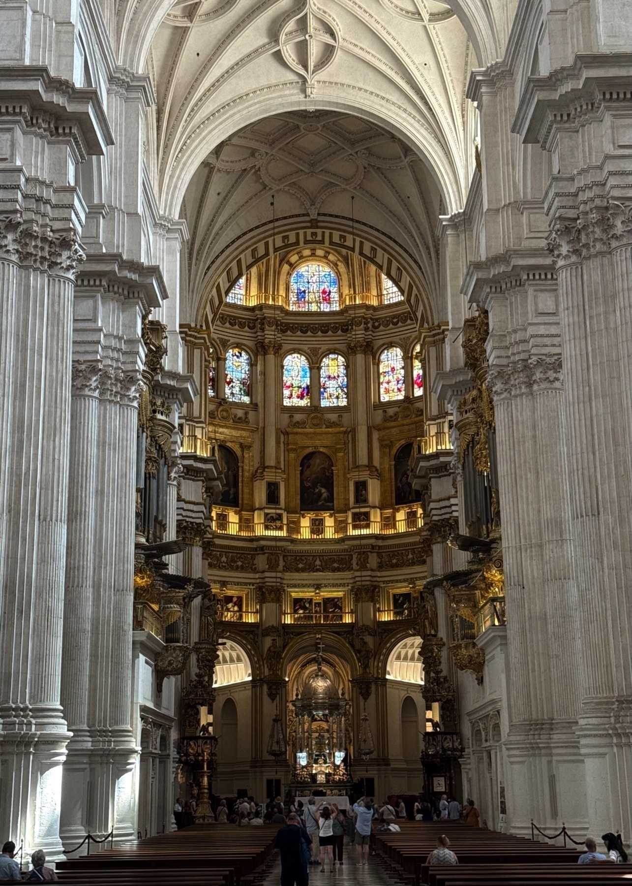 View of the cathedral’s soaring nave leading to the main altar, framed by towering white columns and vaulted ceilings. The richly ornamented apse glows with golden highlights, housing rows of paintings and stained-glass windows that bathe the interior in multicolored light. Visitors walk quietly among pews, admiring the architectural grandeur and spiritual ambiance.