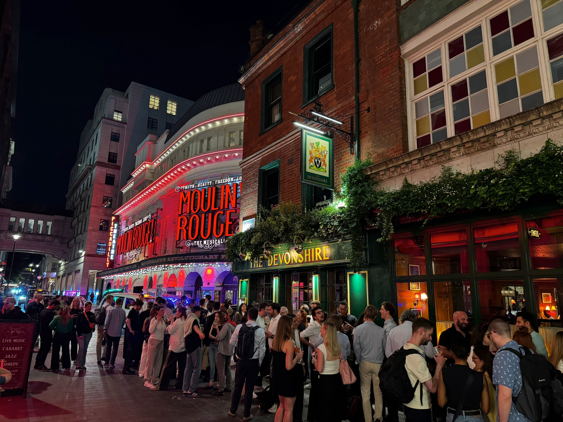 A long evening queue spills down the street outside a pub, packed shoulder to shoulder with people chatting and waiting, while bright red Moulin Rouge theatre lights glow across the road and reflect off the pavement.