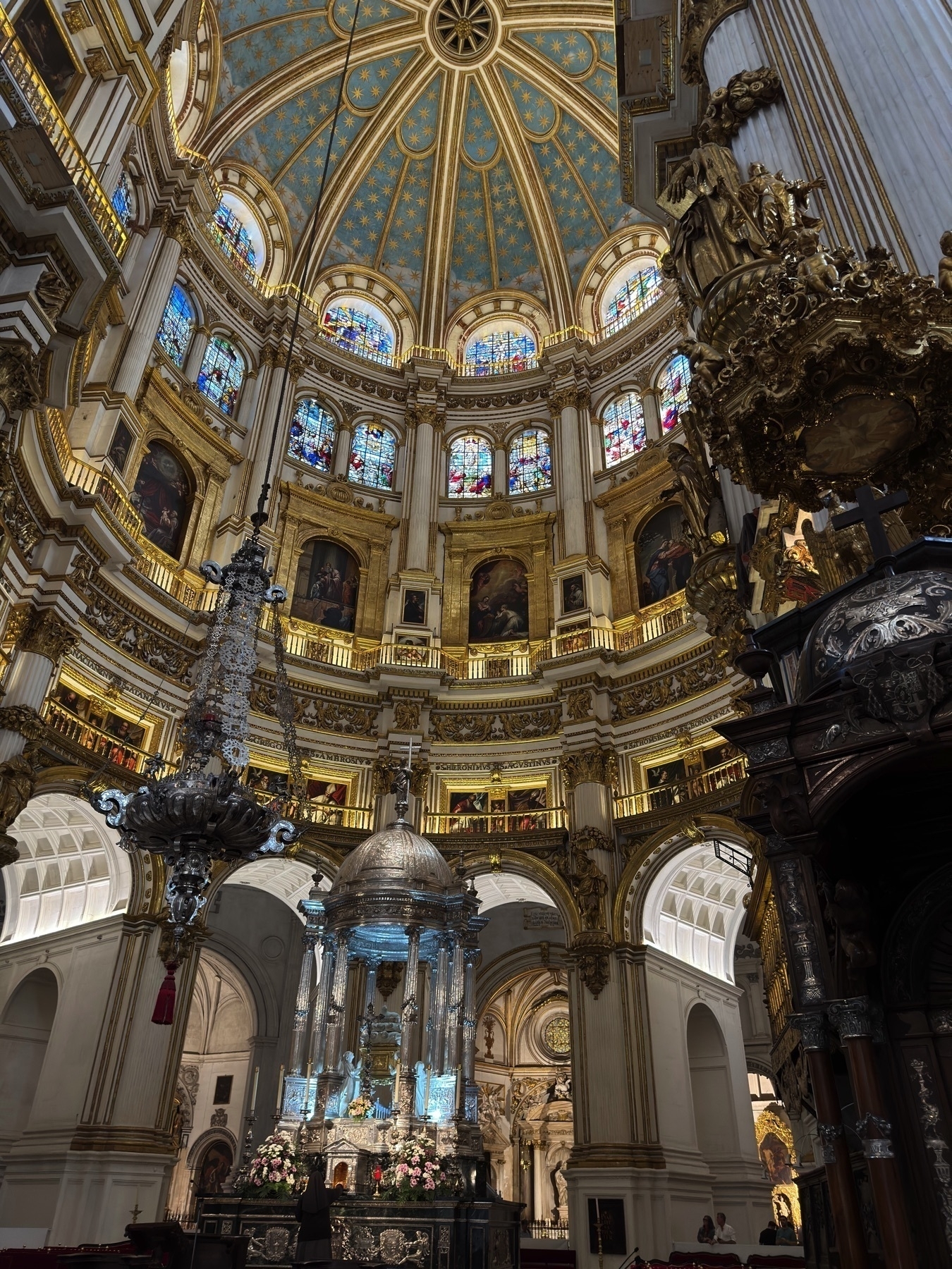 Dramatic upward view of the cathedral’s domed sanctuary, crowned with a celestial blue ceiling adorned with golden stars. Ornate gold detailing and stained-glass windows encircle the dome, while a silver altarpiece shimmers below, surrounded by fresh pink flowers. The composition draws the eye upward, showcasing the majestic verticality and intricate beauty of the space.