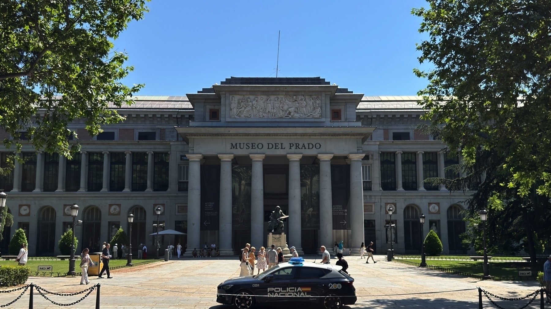 Front view of the Museo del Prado’s main entrance, featuring neoclassical columns, a frieze with sculpted figures, and the museum name in capital letters above the portico. A group of visitors stands in front of the entrance, while a Policia Nacional car is parked in the foreground. Tall trees partially frame the scene on both sides under a clear blue sky.