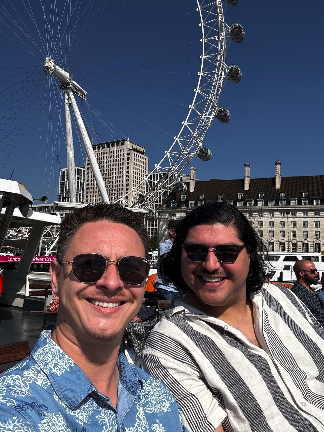Two men smiling on a sunny day aboard a boat, with the towering London Eye Ferris wheel dominating the background. The clear blue sky and vibrant sunlight highlight the details of the wheel and surrounding buildings.
