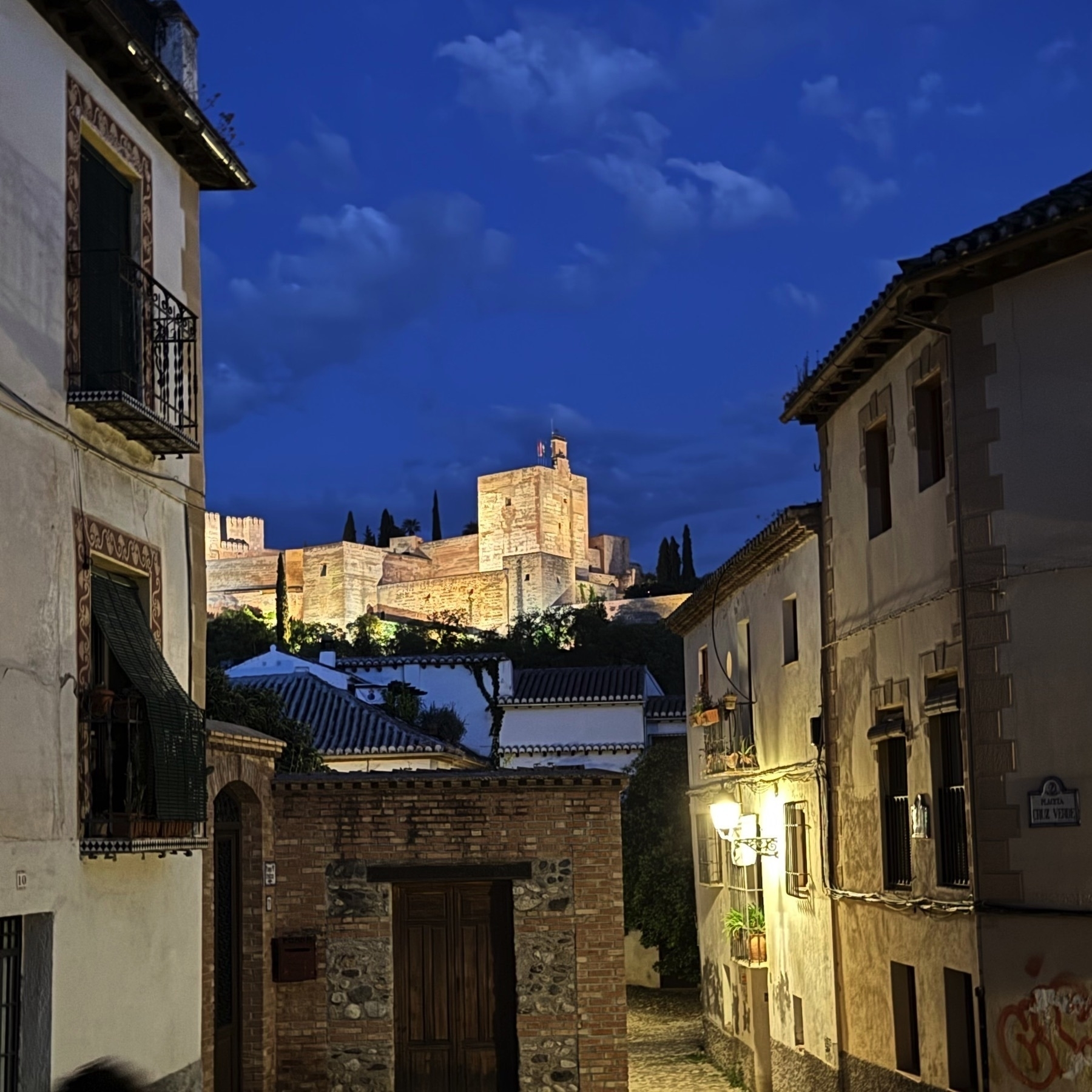 Framed nighttime view of the Alhambra glowing in the distance, seen between traditional Andalusian buildings with tile roofs and wooden balconies. Warm street lighting and the deep blue sky create a peaceful, atmospheric scene.
