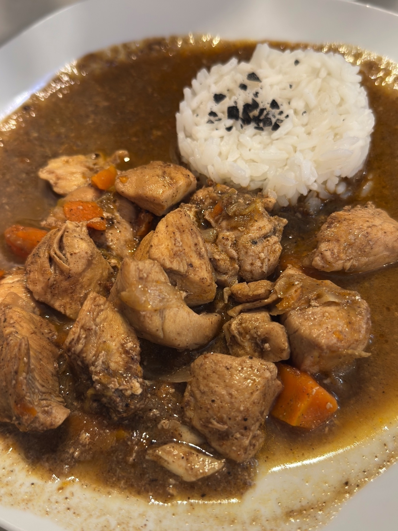 Pollo en curry rojo served in a wide bowl, featuring tender chunks of chicken stewed in a spiced red curry sauce with carrots and onions. A molded dome of white rice topped with black sea salt flakes provides a clean contrast to the deep, glossy broth, creating a rich and comforting presentation.