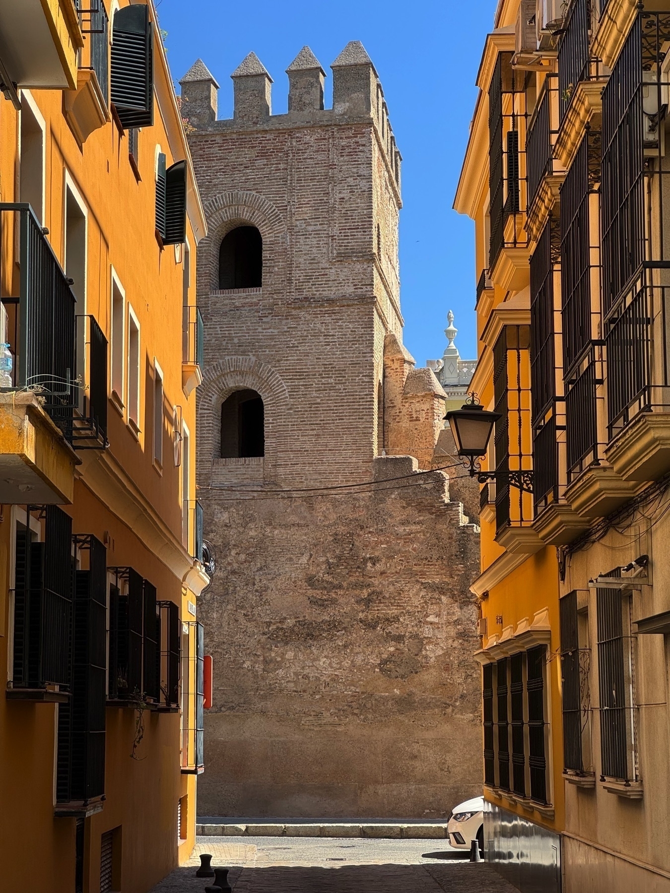 A narrow alley frames a view of a historic brick tower with arched openings and battlements. The sunlit tower contrasts with the colorful yellow and ochre buildings that line the passage, adding depth and warmth to the scene.