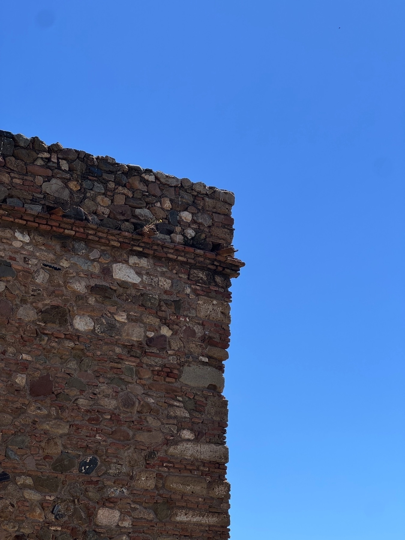 Corner of a weathered stone wall built from a mix of stones and bricks, shown against a deep blue, cloudless sky. The aged texture and rustic construction suggest historical significance, evoking the character of ancient fortifications.