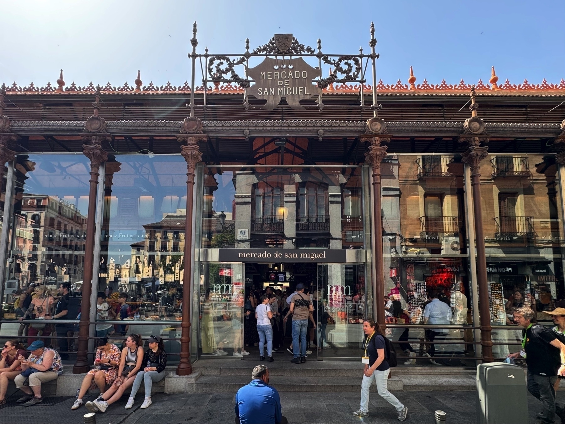 The exterior of Mercado de San Miguel, a historic iron-and-glass market building with ornate architectural details, large glass panels, and decorative wrought-iron signage. The reflections on the glass showcase the surrounding plaza, while a crowd of people gather outside—some sitting on the low railing, others walking or entering through the central doorway.