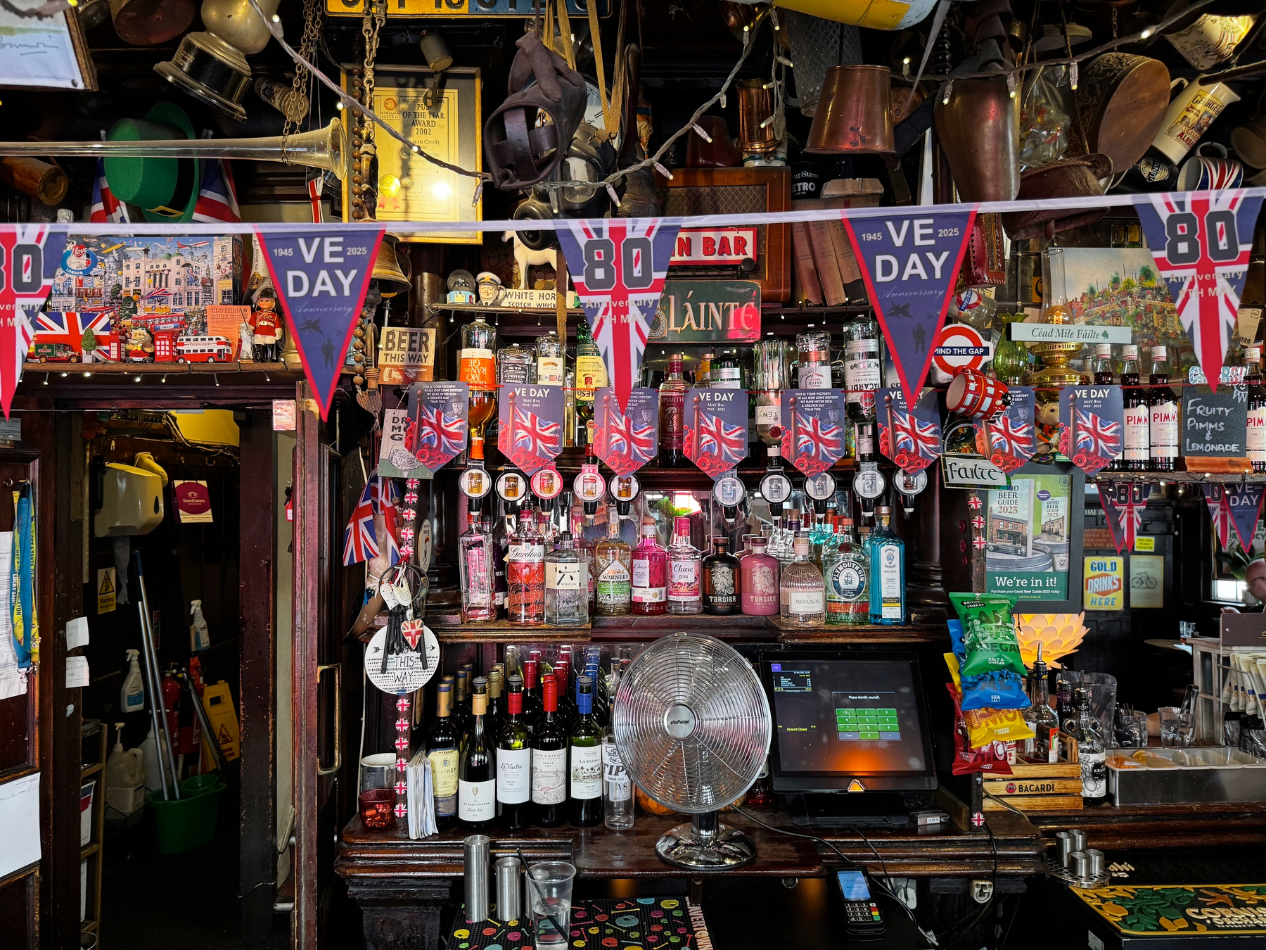 Crowded bar back filled with spirits, taps, and vintage objects, decorated with VE Day 80 bunting and Union Jack flags, creating a dense, celebratory, and historic atmosphere.