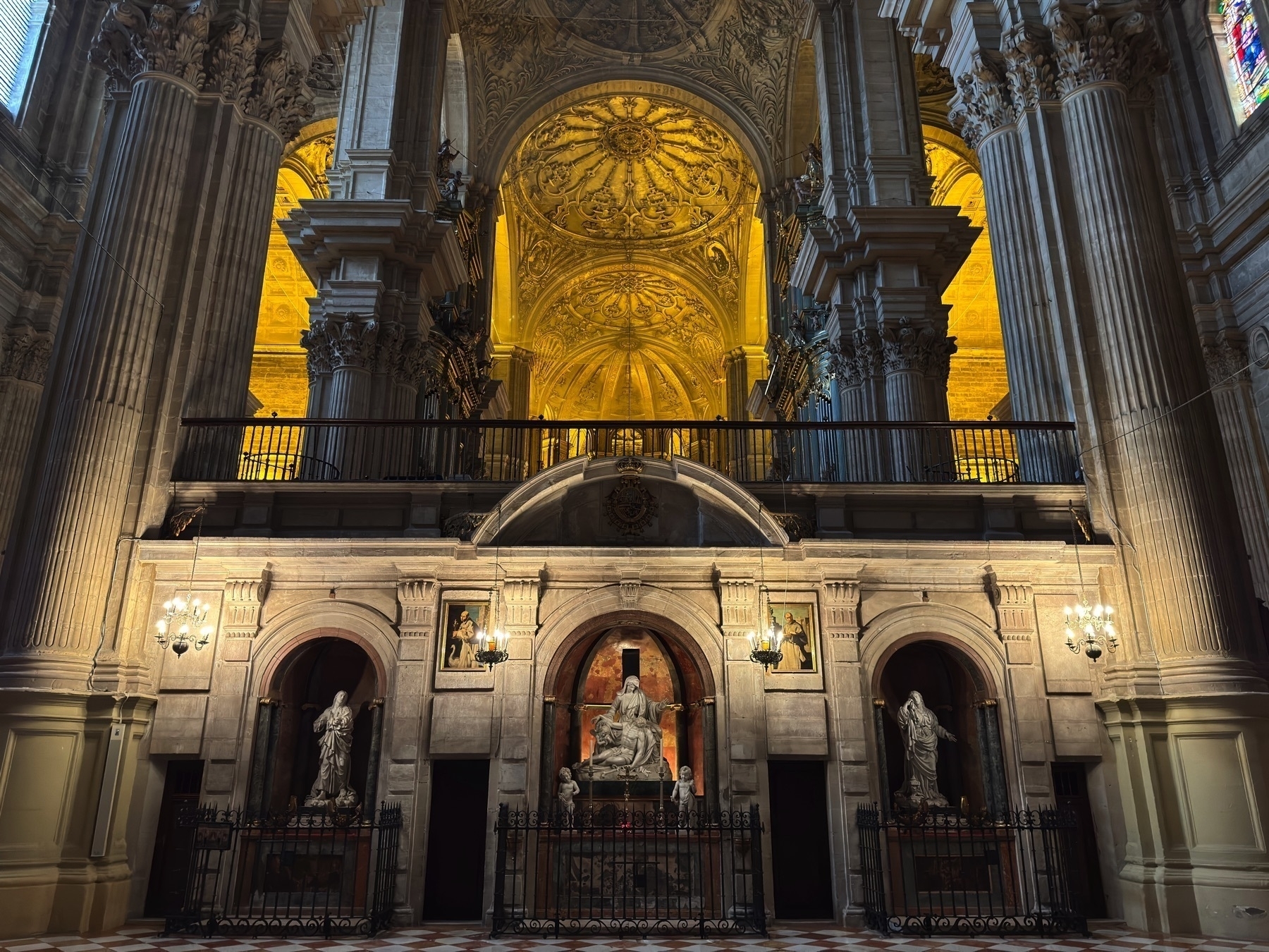A sculptural ensemble of religious figures in stone niches along a wall, framed by massive gray columns and elaborate ceiling vaults above. Warm lighting highlights the detailed carvings and gives the stonework a luminous quality.