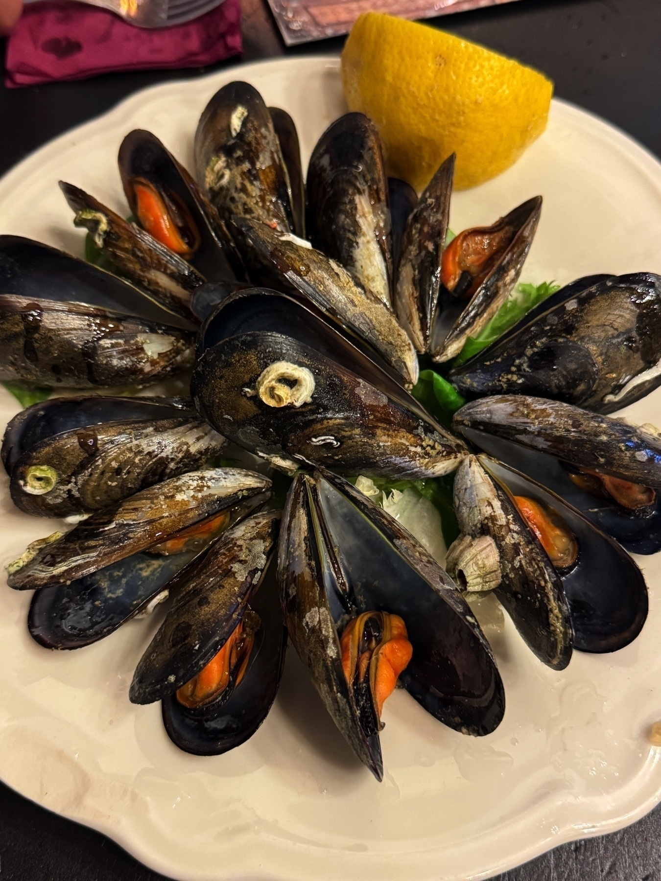 Platter of steamed black mussels arranged in a circular pattern, centered around a wedge of lemon, served on a bed of greens, highlighting the fresh and briny appeal of the seafood.