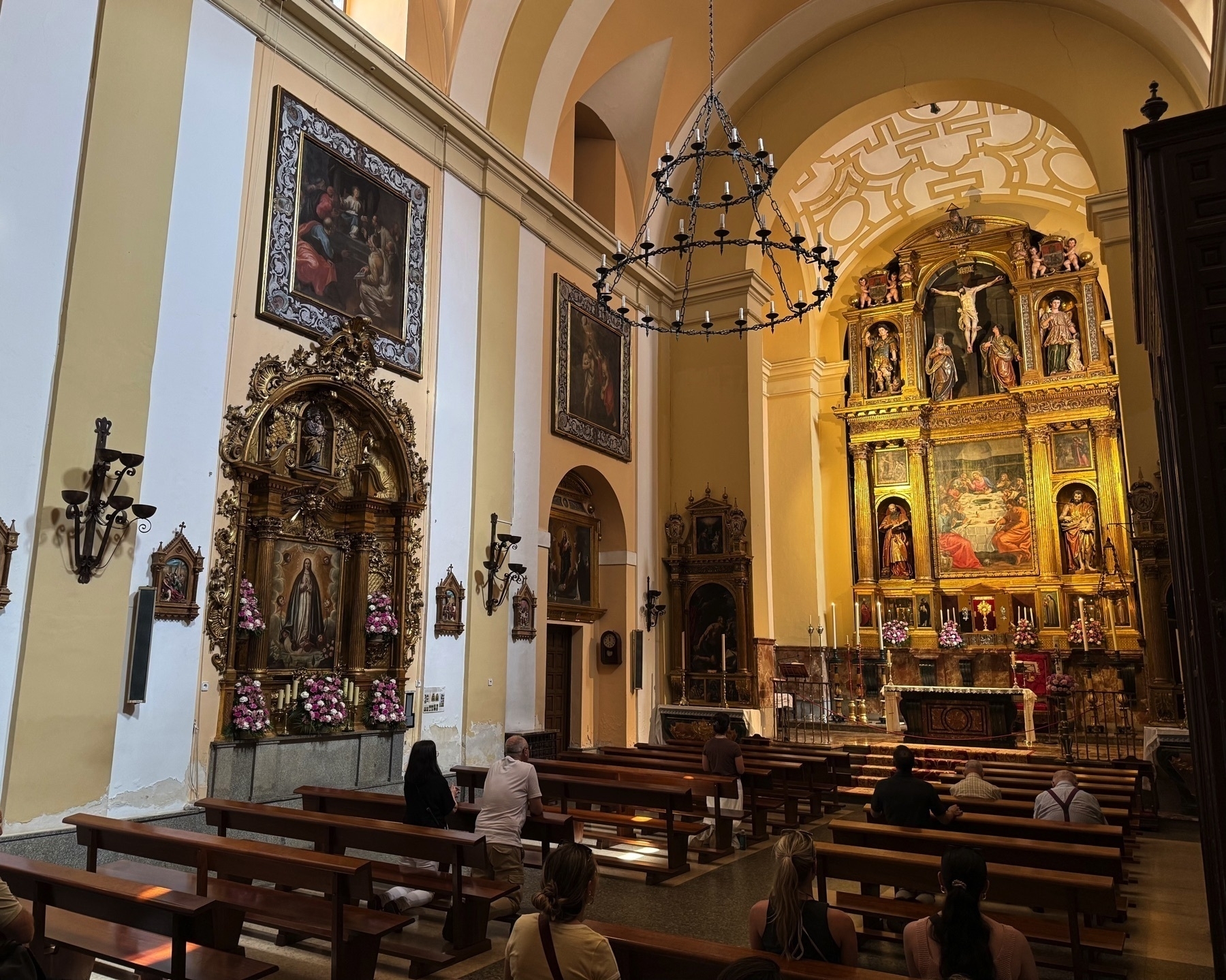 Interior of a richly decorated chapel featuring an elaborate golden altarpiece with religious statues and paintings. Rows of wooden pews are filled with seated visitors facing the altar. The warm yellow walls, arched ceilings, and framed religious art contribute to a peaceful and reverent atmosphere.