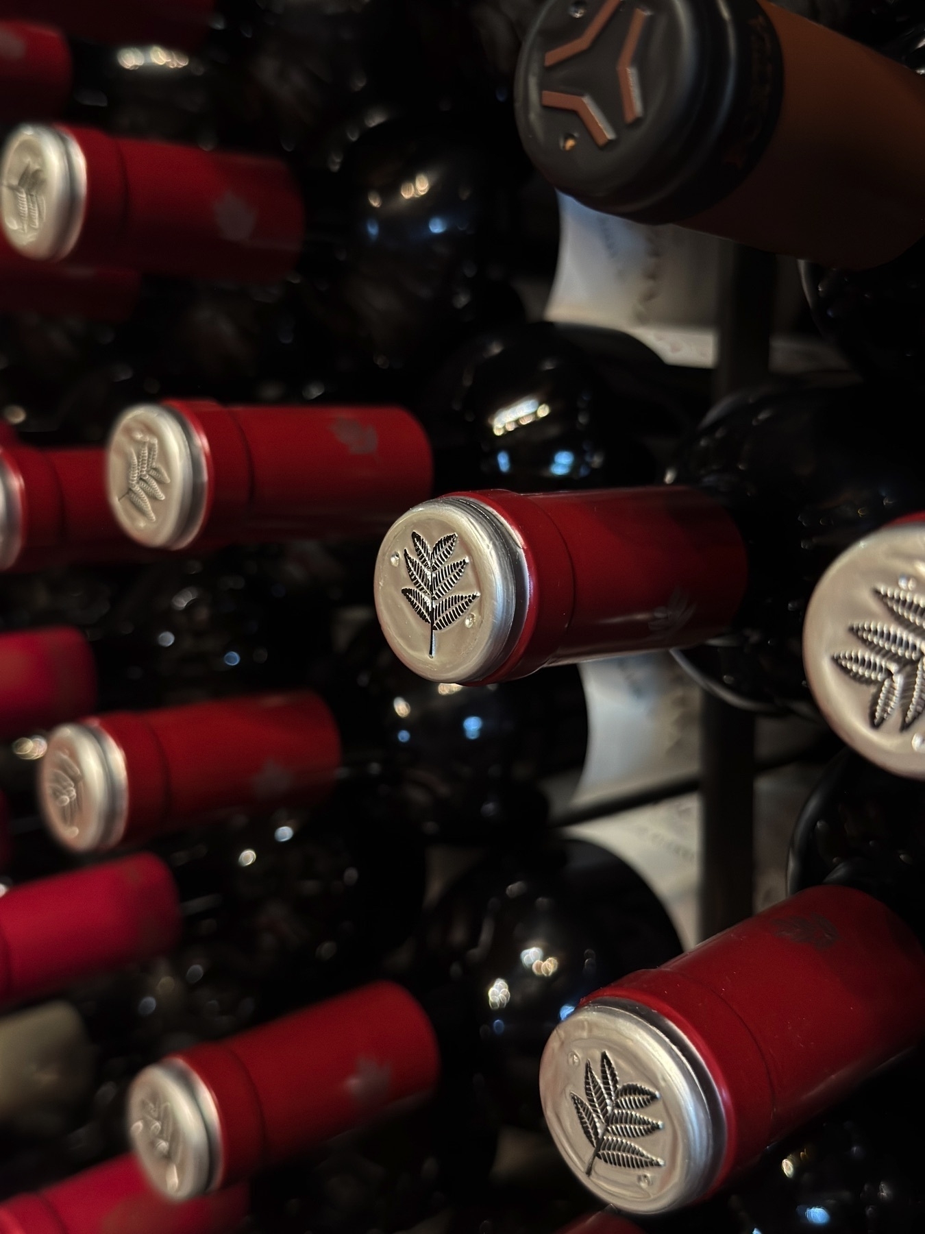 An angled view of a wine rack filled with bottles stacked horizontally, each sealed with a red foil capsule and silver-colored top embossed with a stylized leaf design. The repetitive arrangement and subtle reflections on the dark glass create a visually elegant and orderly composition.