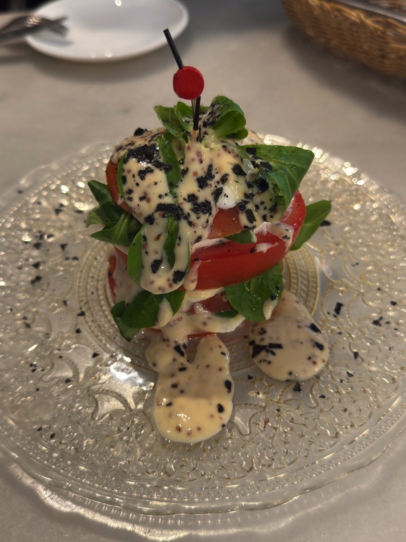 Ensalada de tomate y Mozarella con vinagreta de mostaza y miel, artfully stacked with thick slices of tomato, leafy greens, and rounds of mozzarella cheese. The tower is topped with creamy mustard-honey vinaigrette and speckled with crushed black olives or seeds, served on an ornate clear glass plate that enhances its elegant presentation.