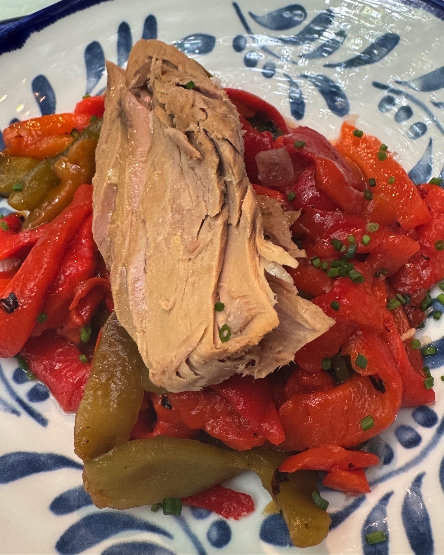 Flaky, light brown fillet of tuna laid over a bed of roasted red and green bell peppers, finely chopped chives sprinkled on top, all served on a ceramic plate with blue decorative patterns.